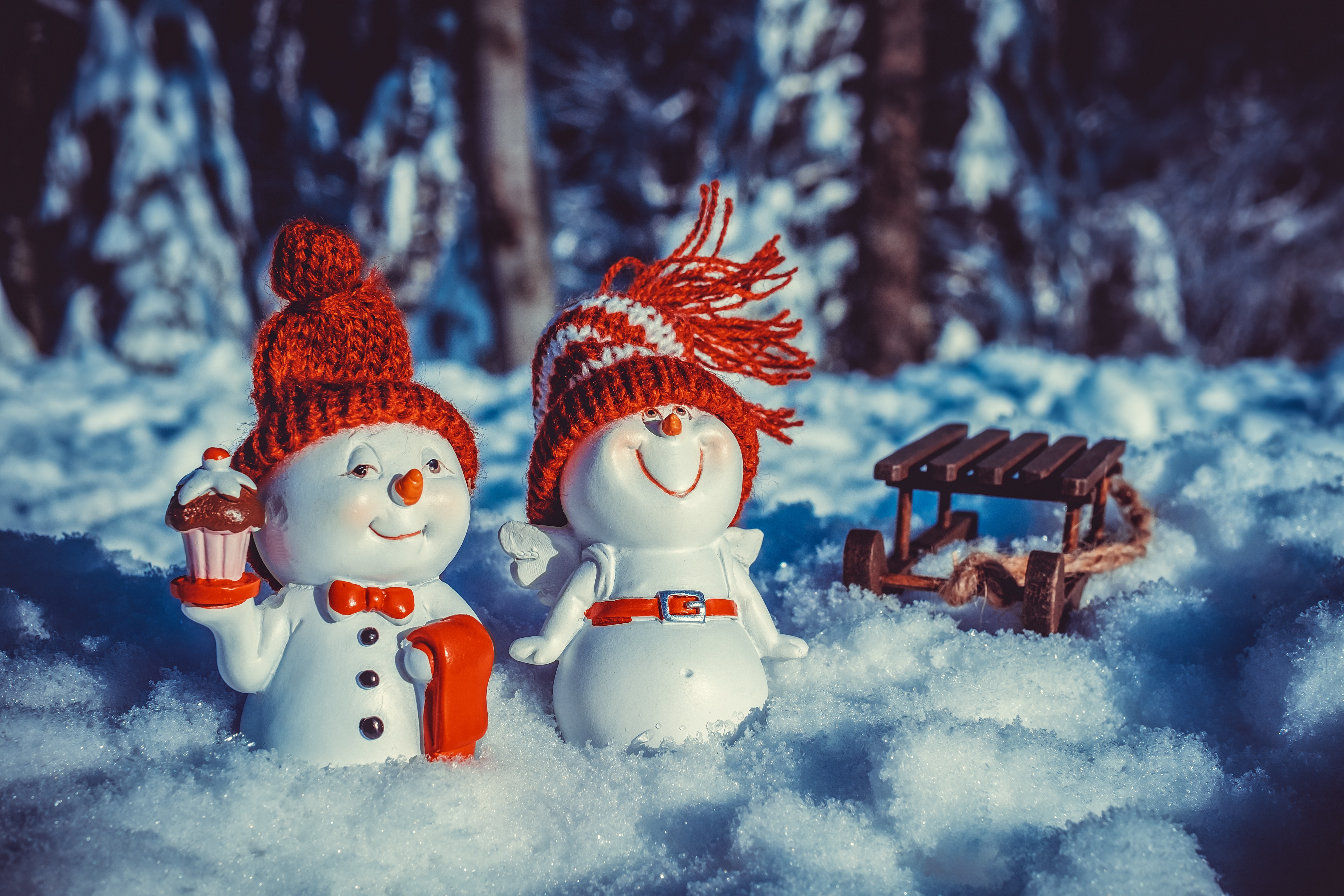 Two adorable snowmen figurines in red and white hats stand in the snow, a miniature wooden sled sits behind them. Festive winter scene perfect for a holiday wallpaper