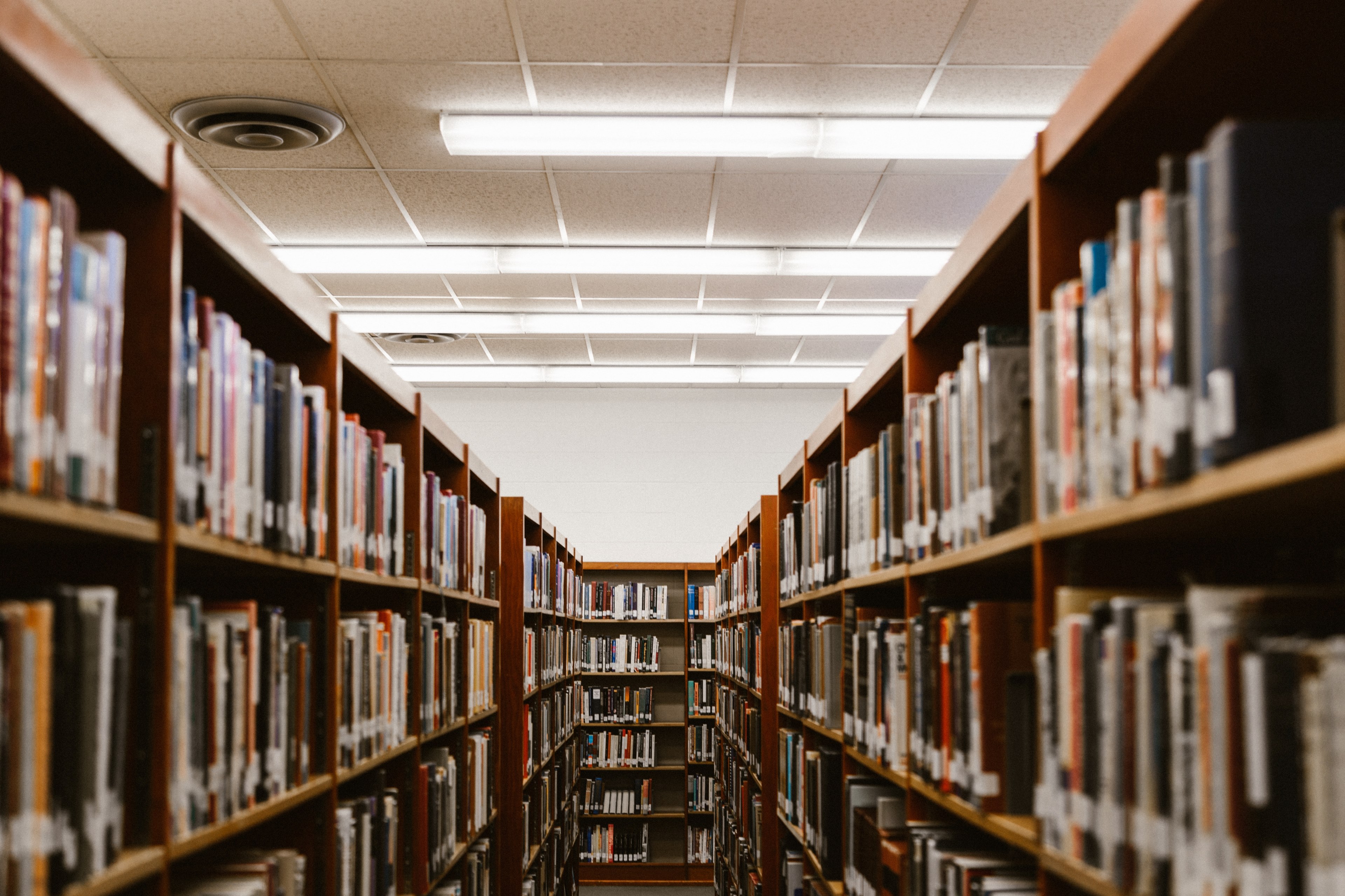 A long, narrow aisle between tall bookshelves filled with various books in a library. The warm, wooden shelves contrast with the bright fluorescent lights illuminating the space, creating a cozy atmosphere for learning and discovery
