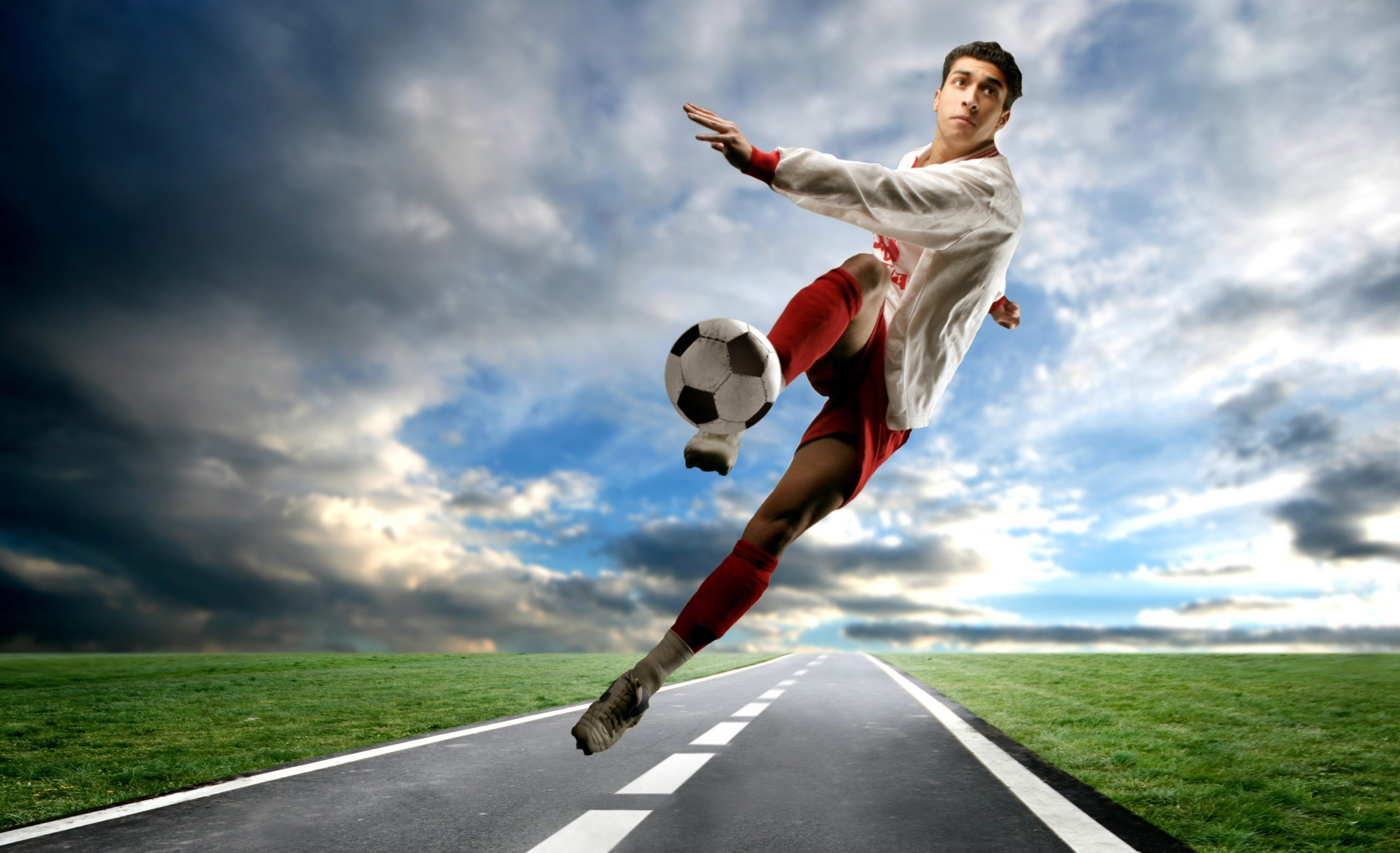A soccer player dynamically kicks the ball in mid-air above a paved road, against a dramatic sky backdrop