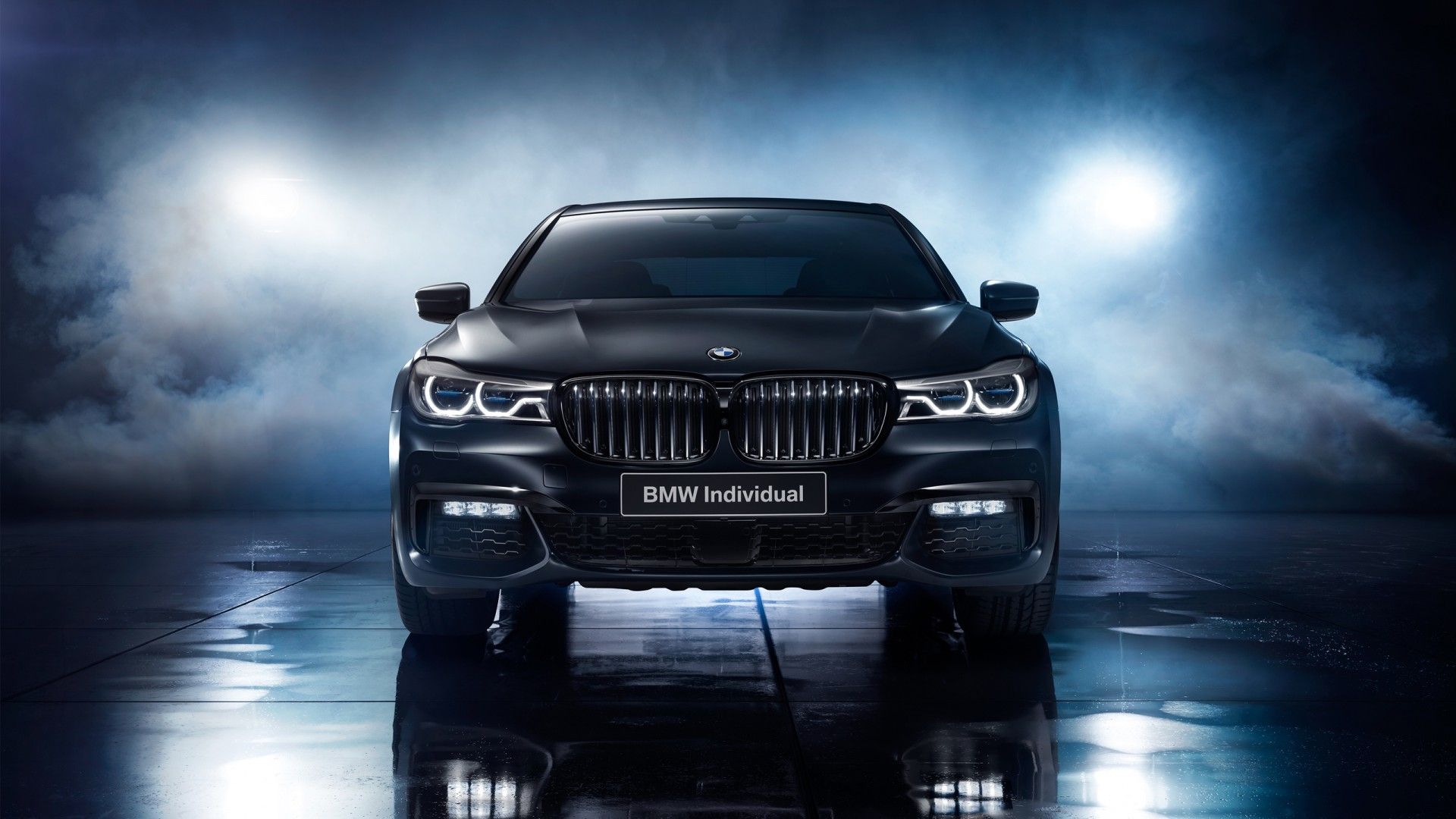 A front view of a sleek, black BMW Individual 7 Series sedan, illuminated by headlights, against a dark, smoky backdrop