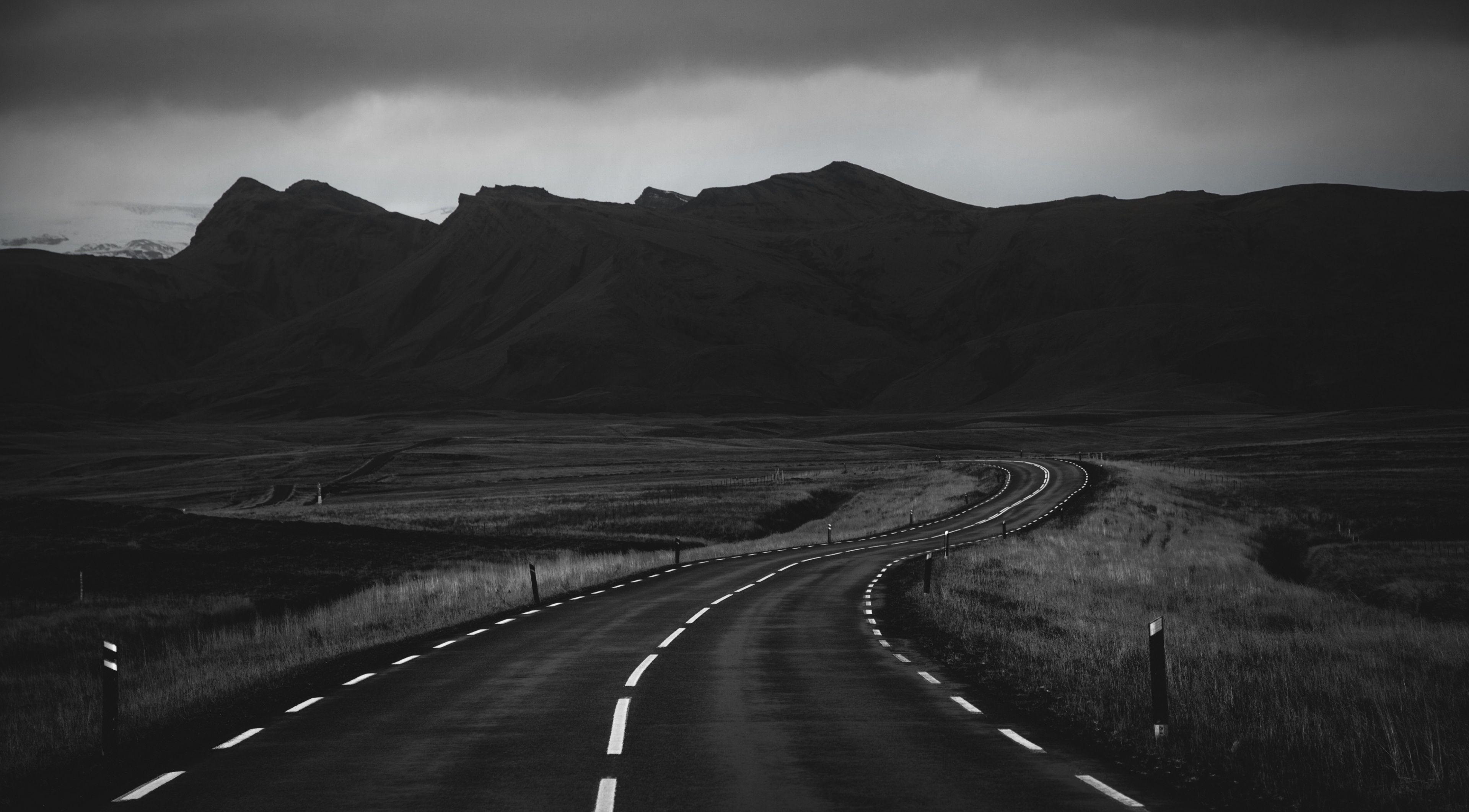 A grayscale photograph of a winding road leading towards dark, imposing mountains under a brooding, cloudy sky