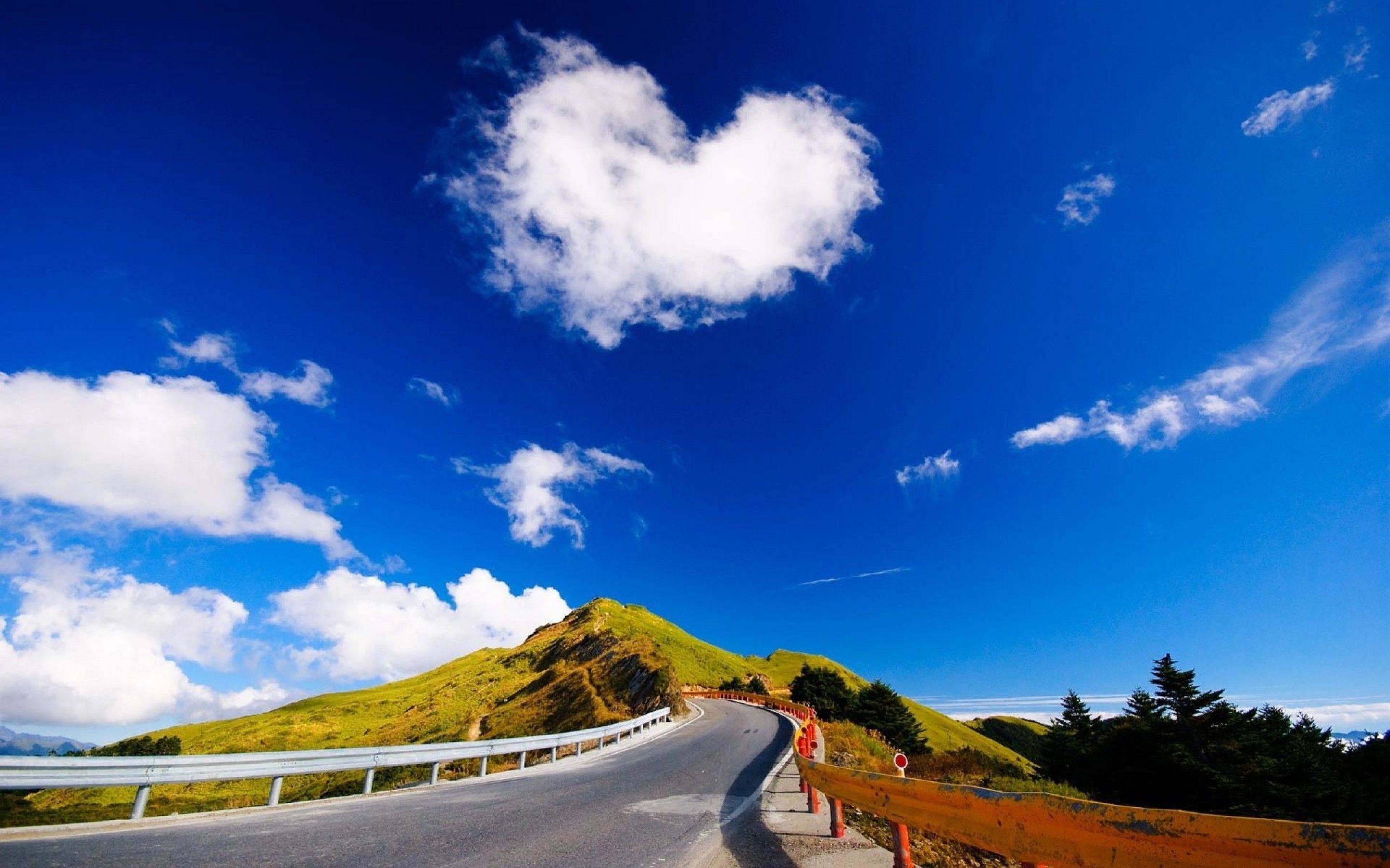 A scenic road winds its way up a green mountain beneath a heart-shaped cloud and a vivid blue sky