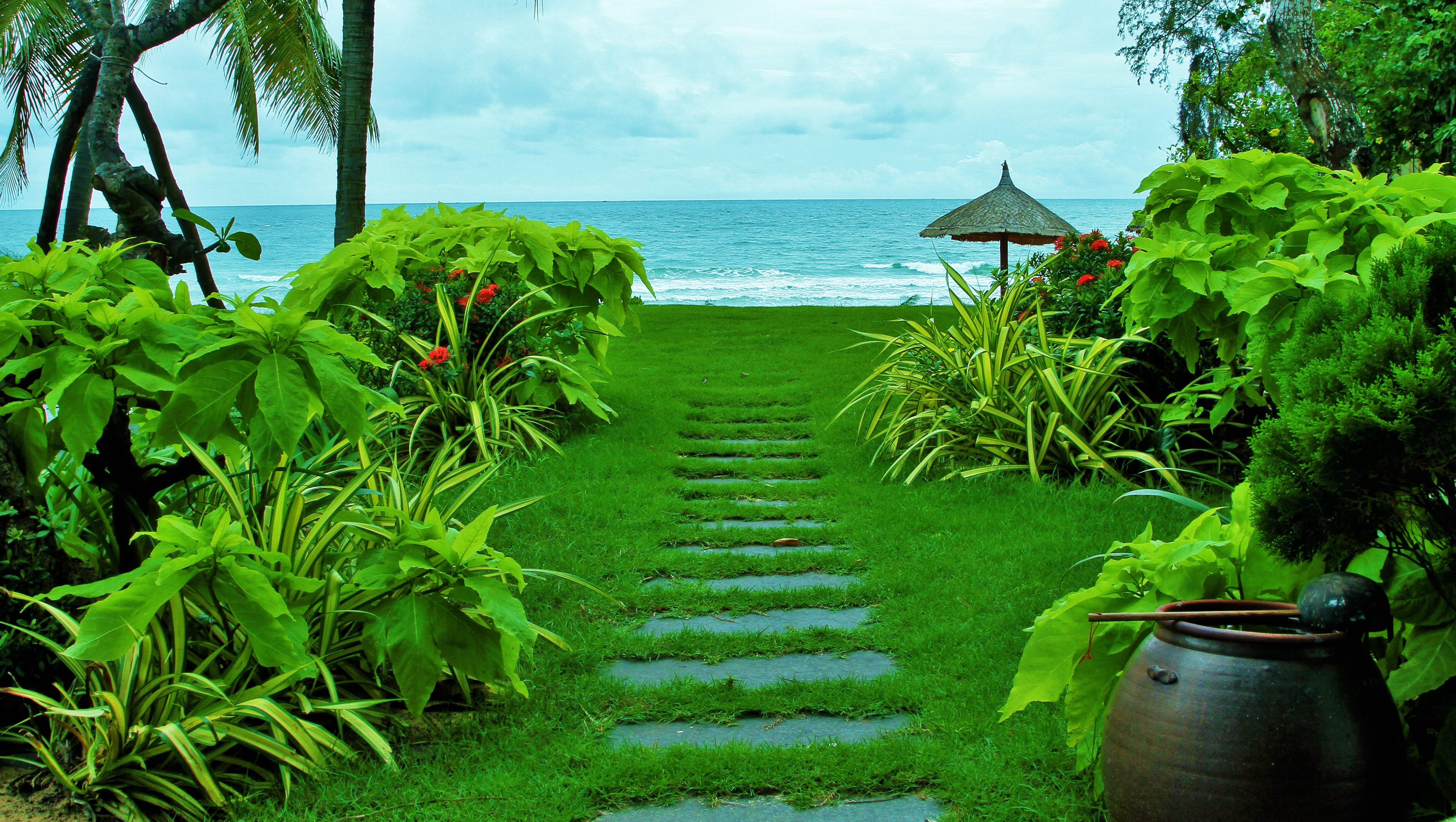 A lush green pathway leading to a serene beach, surrounded by tropical plants and a thatched umbrella