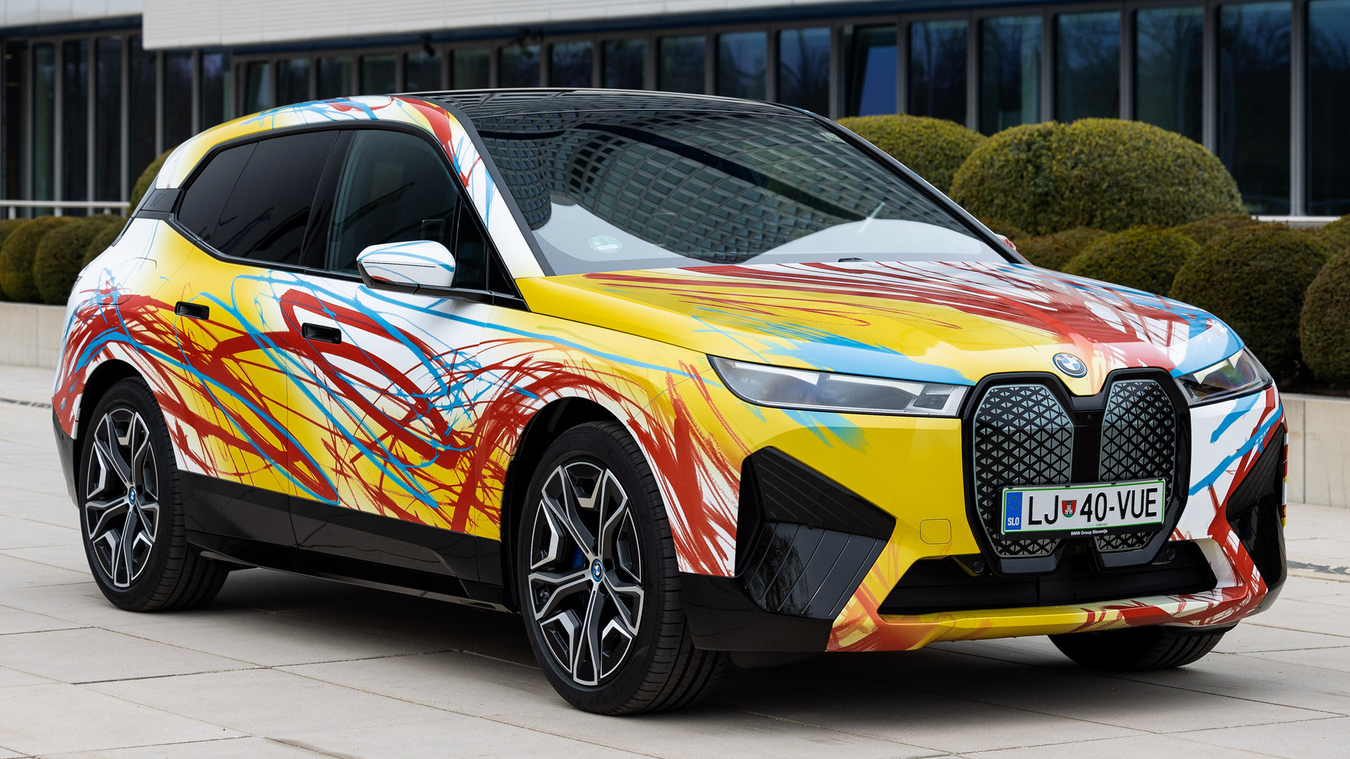 A BMW iX electric SUV with a unique, colorful abstract art design displayed in front of a modern building