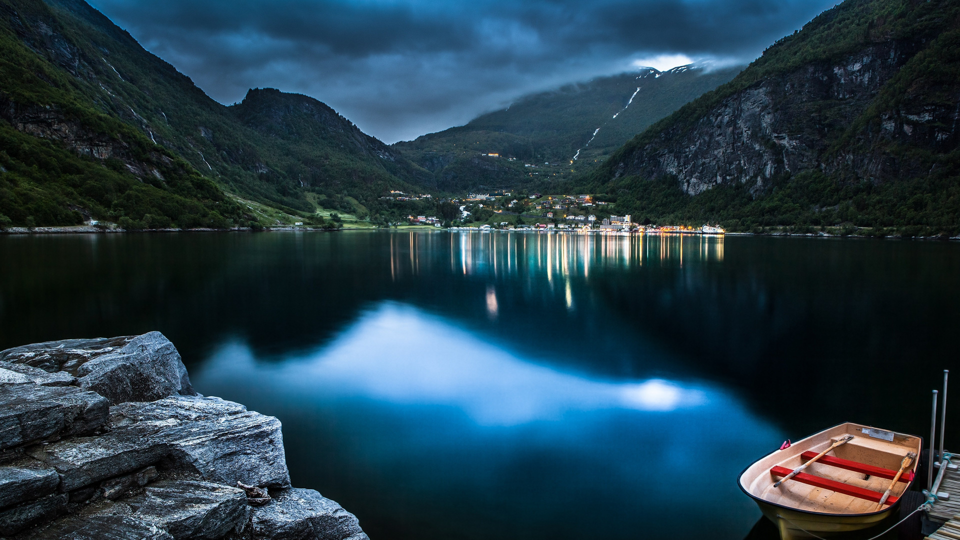 A serene 4k wallpaper of a fjord in Norway at dusk, with a small boat moored in the foreground and the lights of a village reflected in the calm water