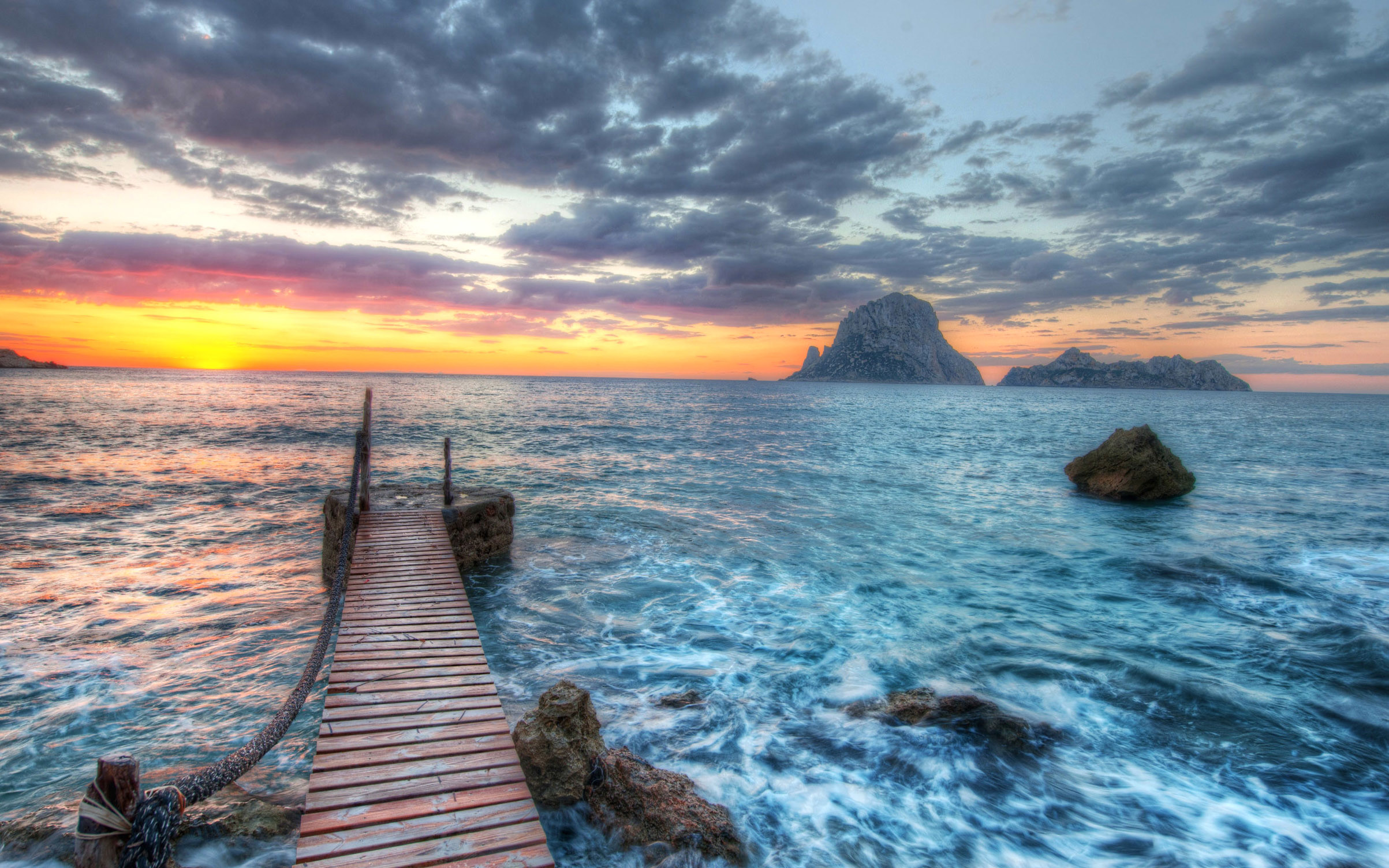 A wooden pier stretches out into the vibrant ocean at sunset, with rocky islands silhouetted against the colorful sky