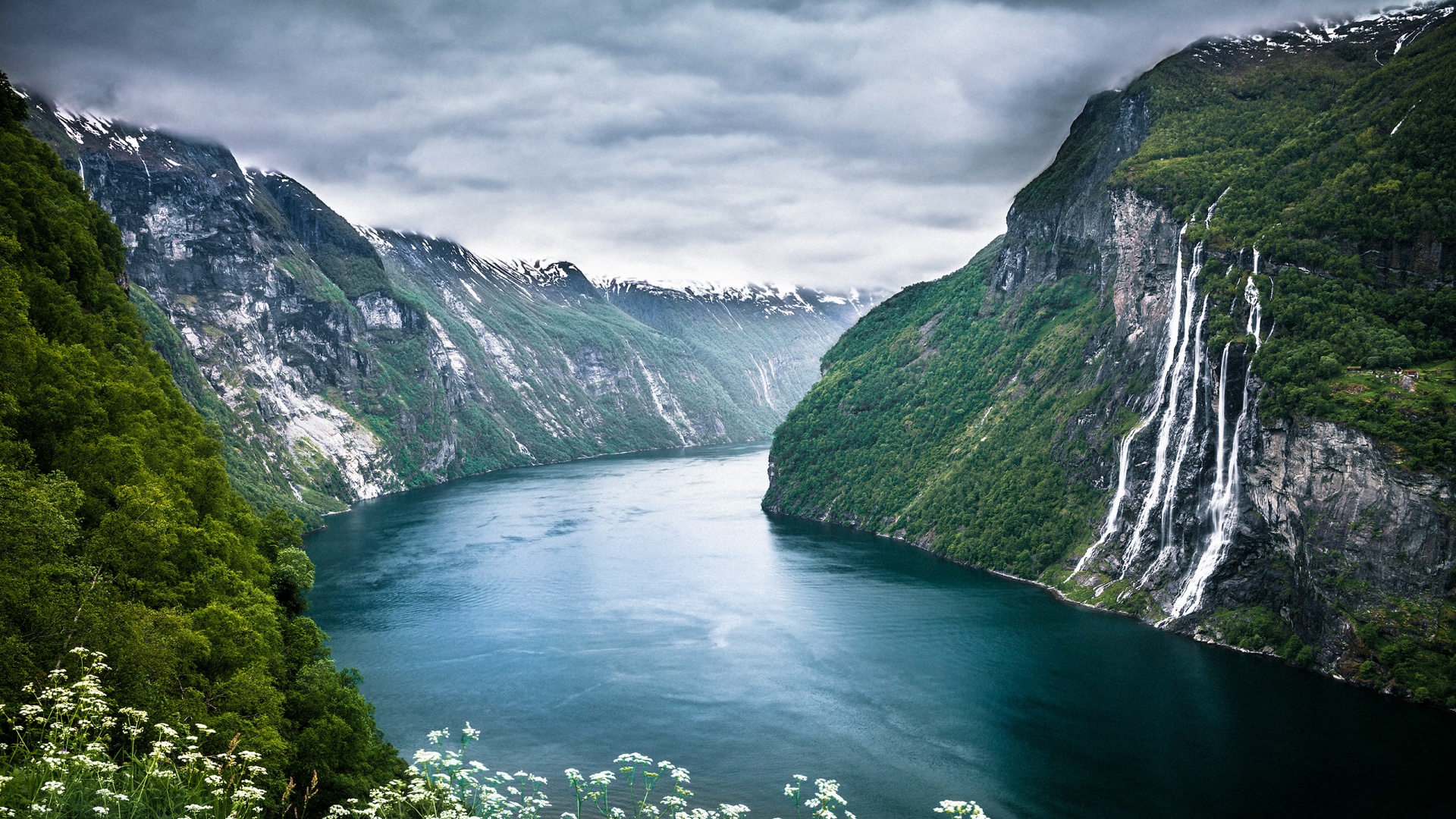 A breathtaking view of a fjord in Norway, with steep, snow-capped mountains flanking the deep blue water. Several waterfalls cascade down the cliffs, adding to the dramatic scenery