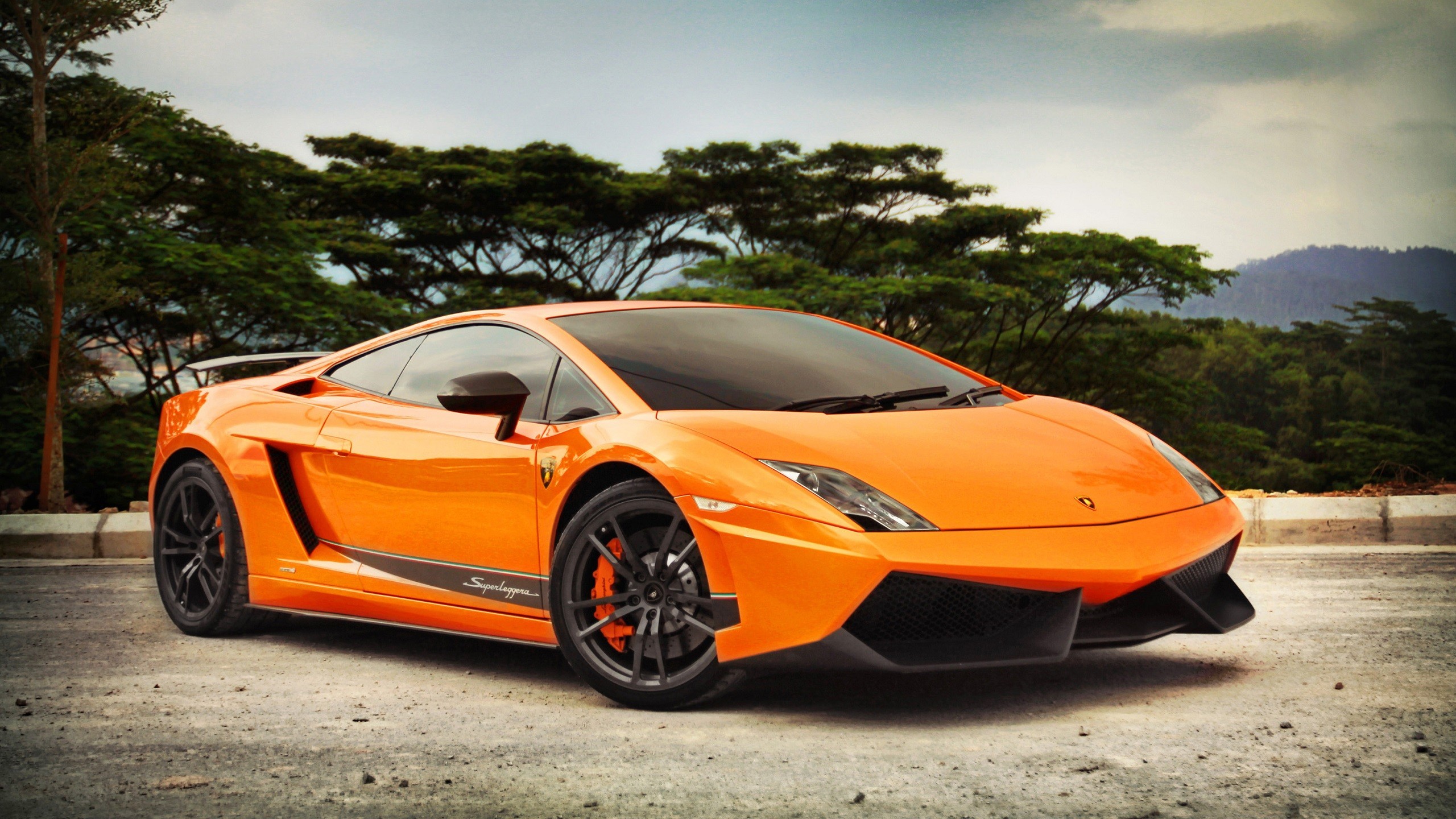 A vibrant orange Lamborghini Gallardo parked on a roadside, with lush green trees and a distant hill in the background