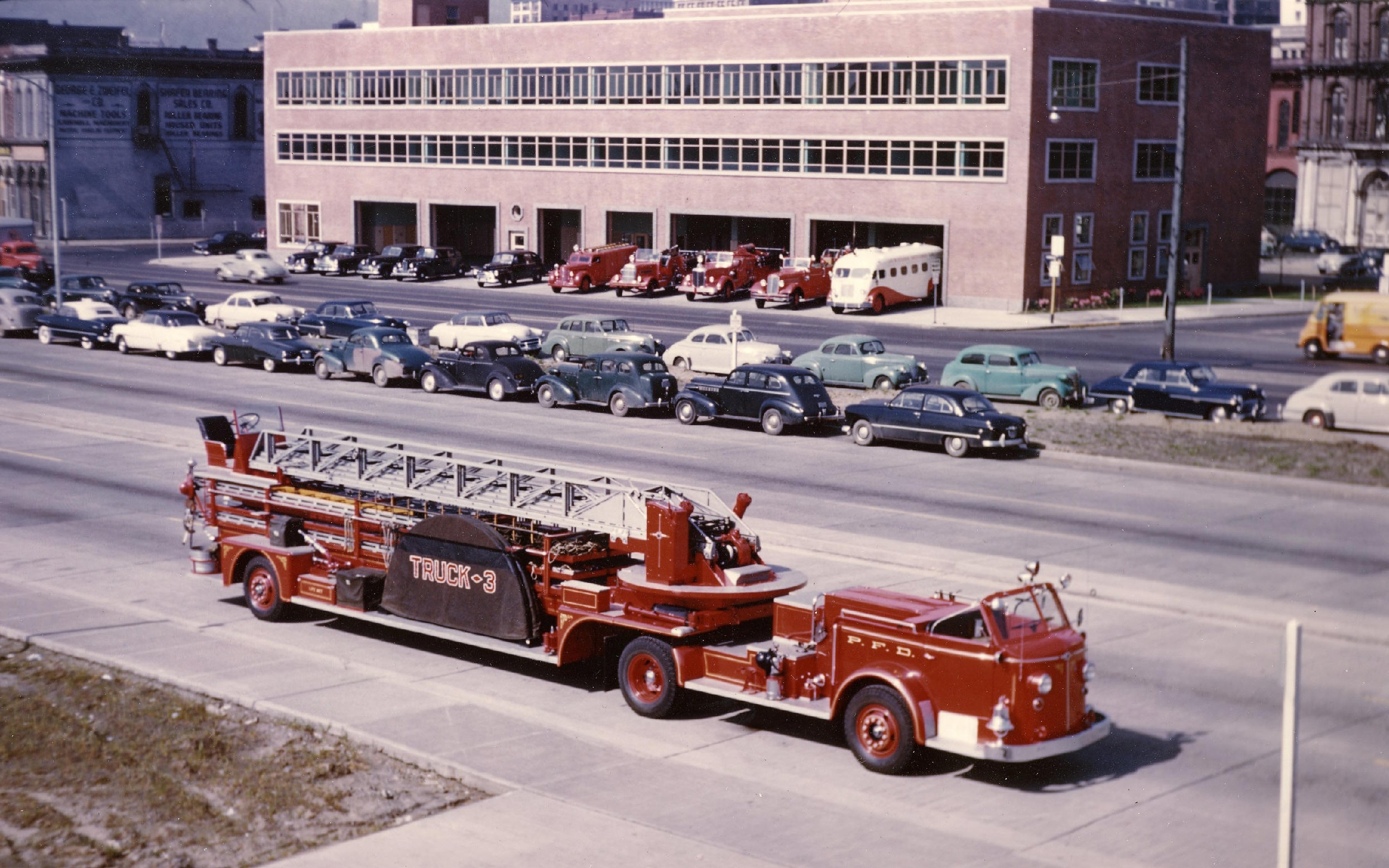 A vintage fire truck, marked 'Truck-3', is prominently displayed in the foreground of the image.  It's a classic red design, with a long extendable ladder. In the background, a street lined with parked cars, various makes and models from the mid-20th century, stretches towards a brick building.  The building appears to be a fire station, with several fire trucks visible within its open bays.  This suggests the image may be a historical record of a fire department or a snapshot of city life from a bygone era