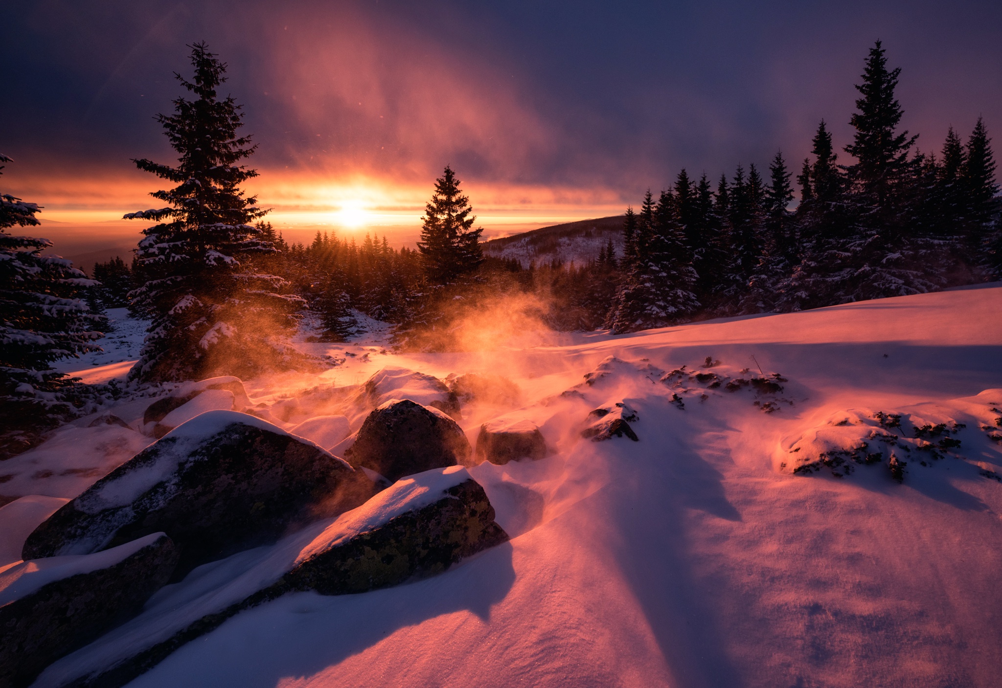 A breathtaking 4k winter wallpaper showcasing a snowy mountain landscape at sunset. The warm hues of the setting sun paint the snow-covered ground and pine trees, creating a stunning contrast against the cool purple and blue tones of the sky. Wisps of snow are gently blown by the wind, adding a touch of magic to the scene
