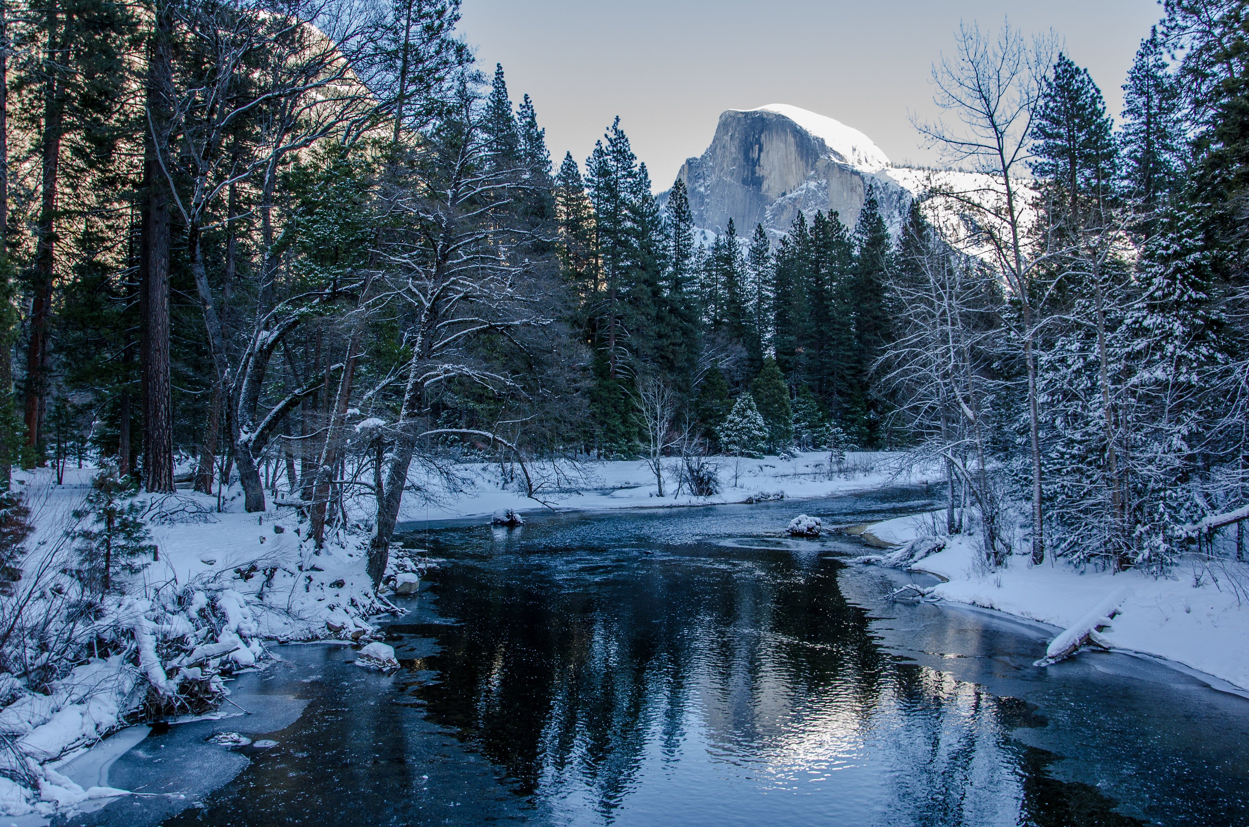 A serene winter landscape featuring a snow-dusted forest, a calm river reflecting the sky, and a majestic mountain in the background
