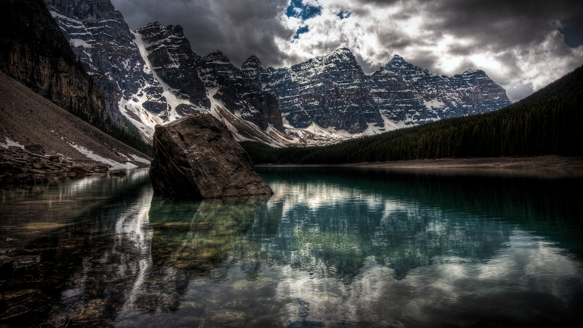 Moraine Lake in Banff National Park with a large rock in the foreground, surrounded by snow-capped mountains and a dark forest under a dramatic cloudy sky
