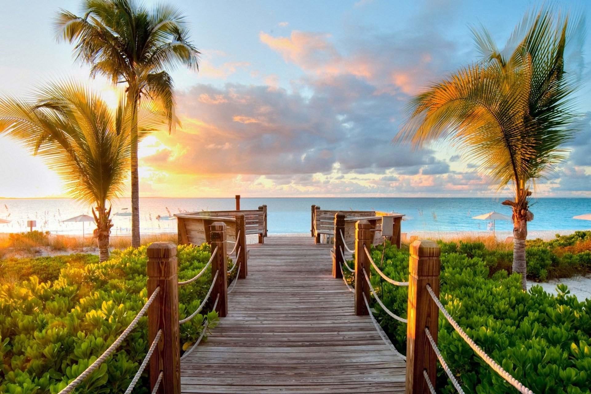 A picturesque wooden boardwalk leading to a tranquil beach at sunset, with palm trees swaying gently in the breeze and calm turquoise waters