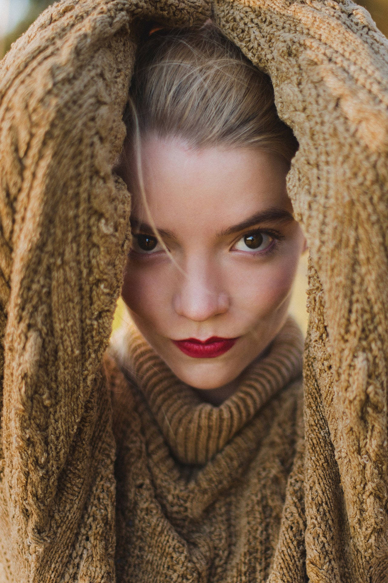 Close-up of Anya Taylor-Joy peering through a brown knitted sweater, her blonde hair pulled back and her red lipstick highlighting her lips