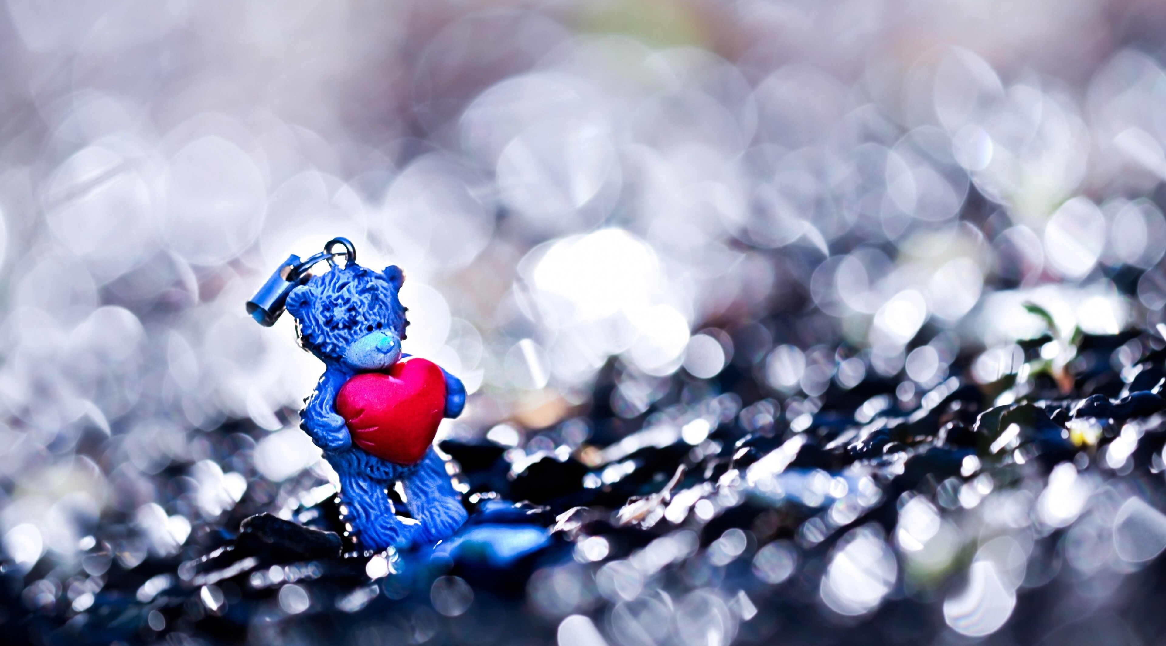 A cute blue teddy bear keychain holds a red heart, symbolizing Valentine's Day love against a blurred, sparkling background