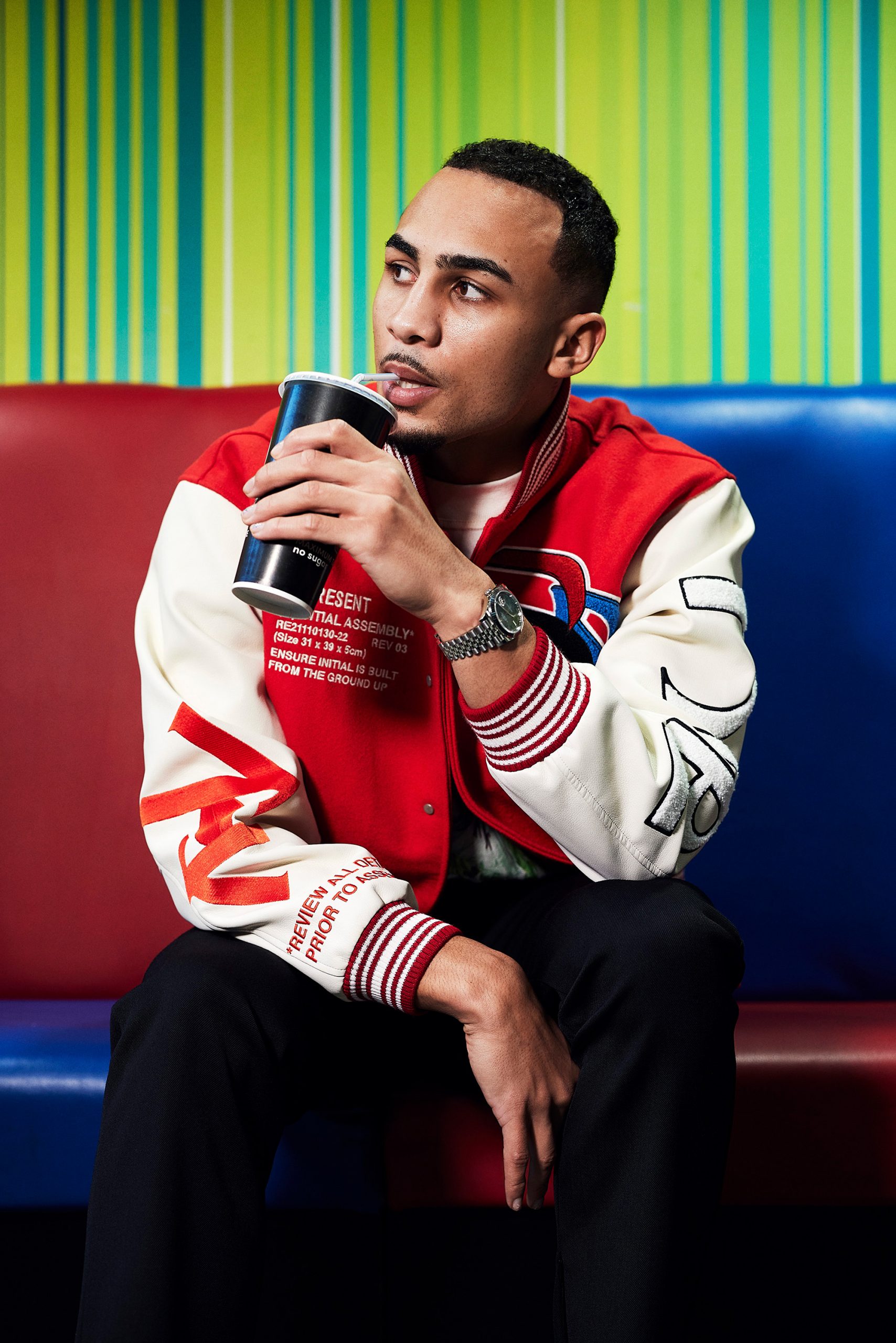 Benjamin Whittaker enjoying a drink while sitting on a vibrant, striped sofa. He is wearing a stylish red and white varsity jacket, paired with dark pants. He looks off-camera with a thoughtful expression