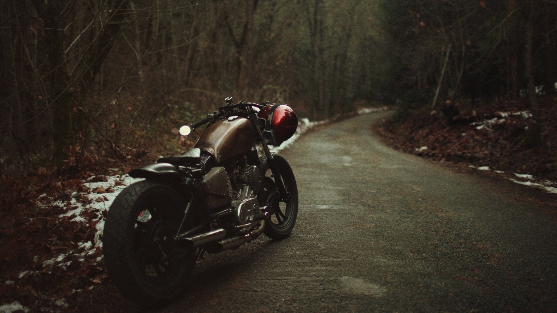 A custom-built motorcycle parked on a quiet road in a wooded area. The bike has a vintage, rugged look and features a dark metallic finish. A red helmet hangs from the handlebars, adding a pop of color to the scene
