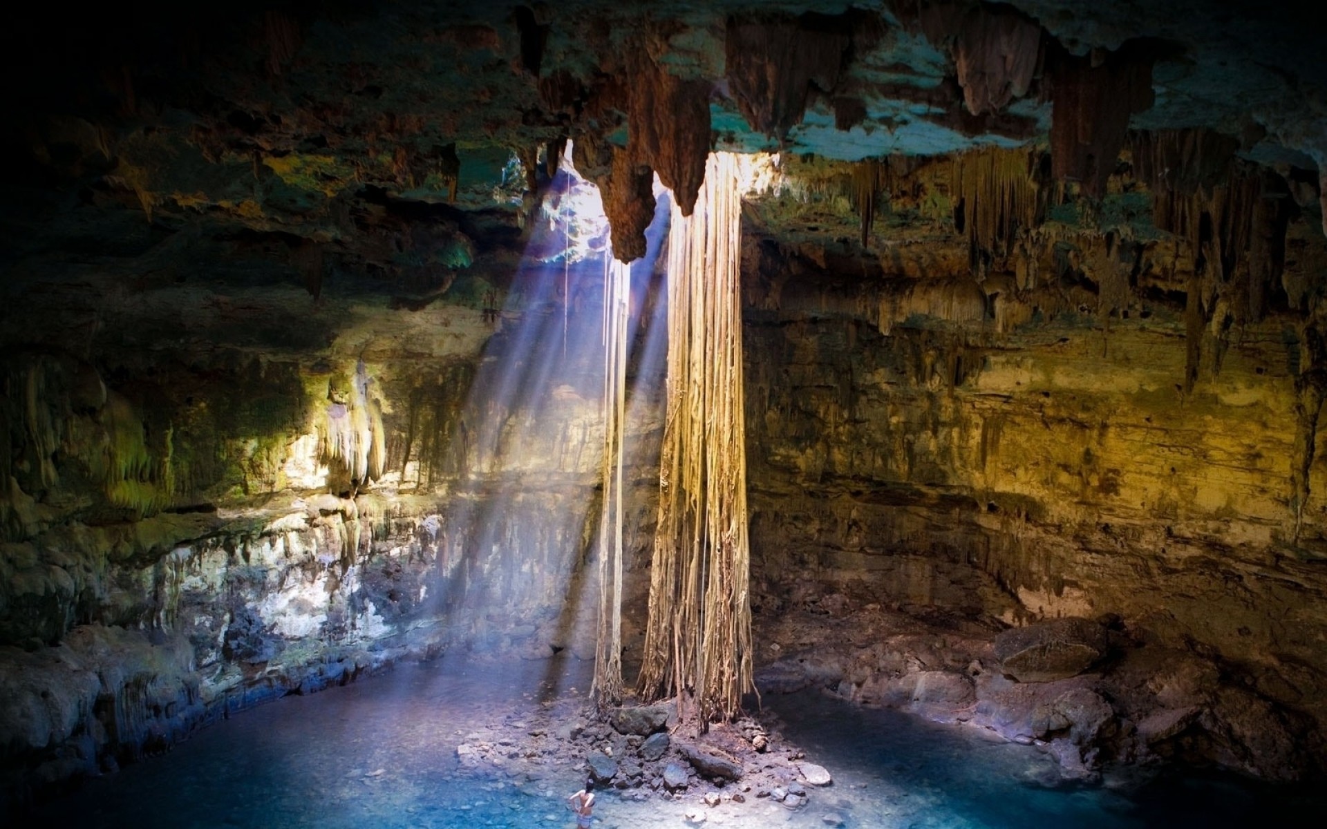 Sunbeams illuminate a cenote in the Yucatan Peninsula, Mexico. Stalactites hang from the ceiling and the water below glows blue