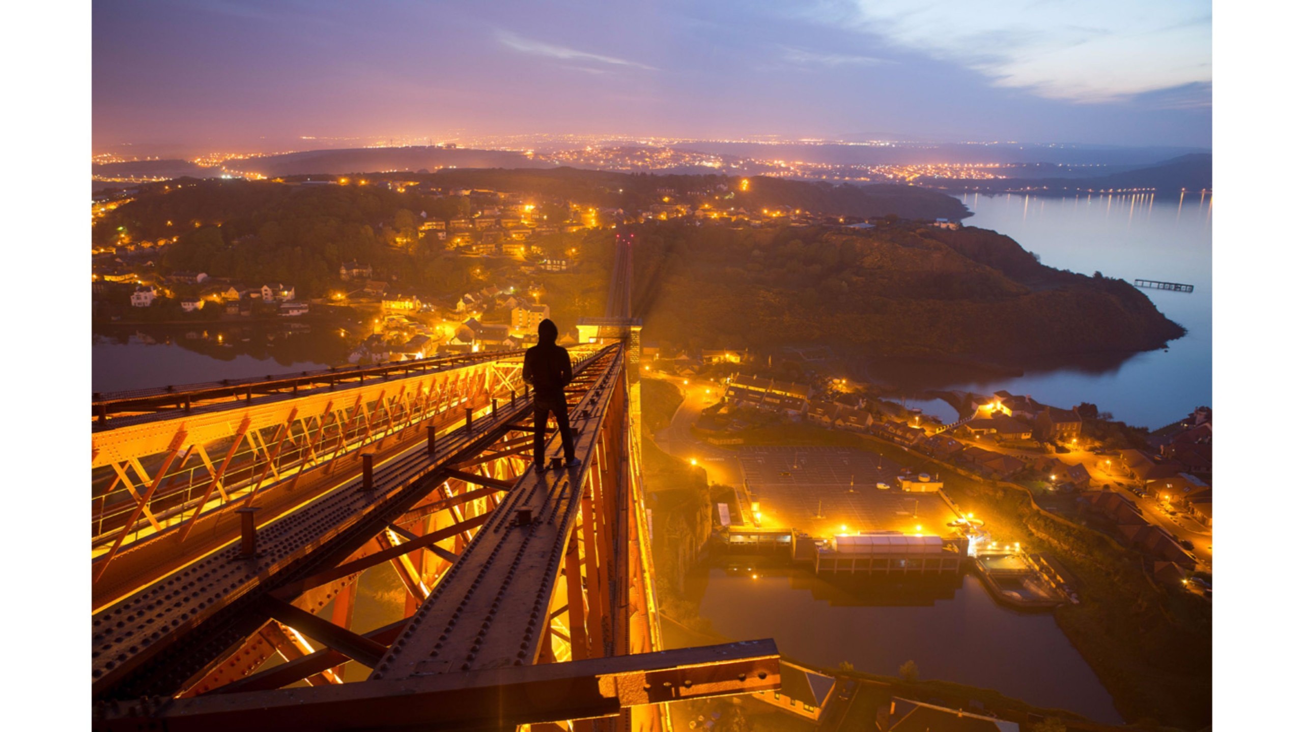 A silhouetted figure stands atop the Forth Rail Bridge at night, overlooking a twinkling cityscape and tranquil waters