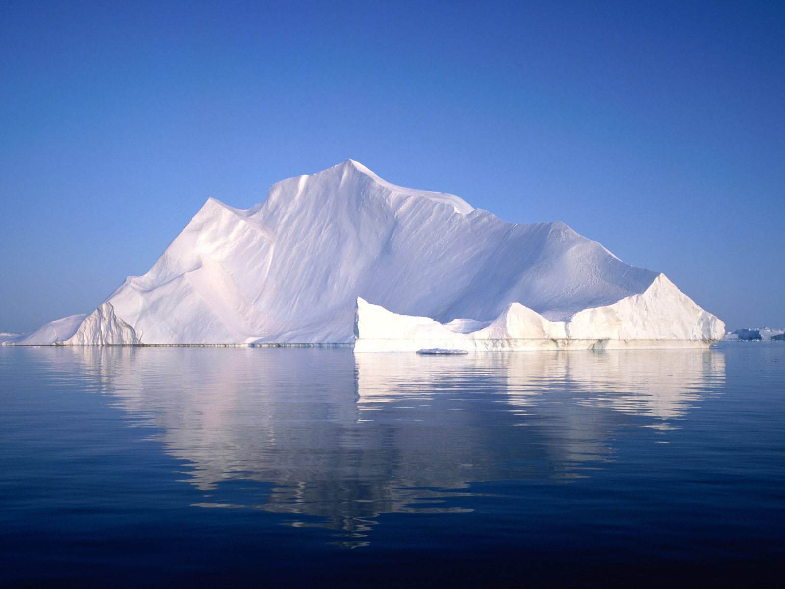A majestic iceberg, mostly white, rises from the dark blue water, its reflection shimmering on the surface. The clear blue sky forms a backdrop to this icy giant