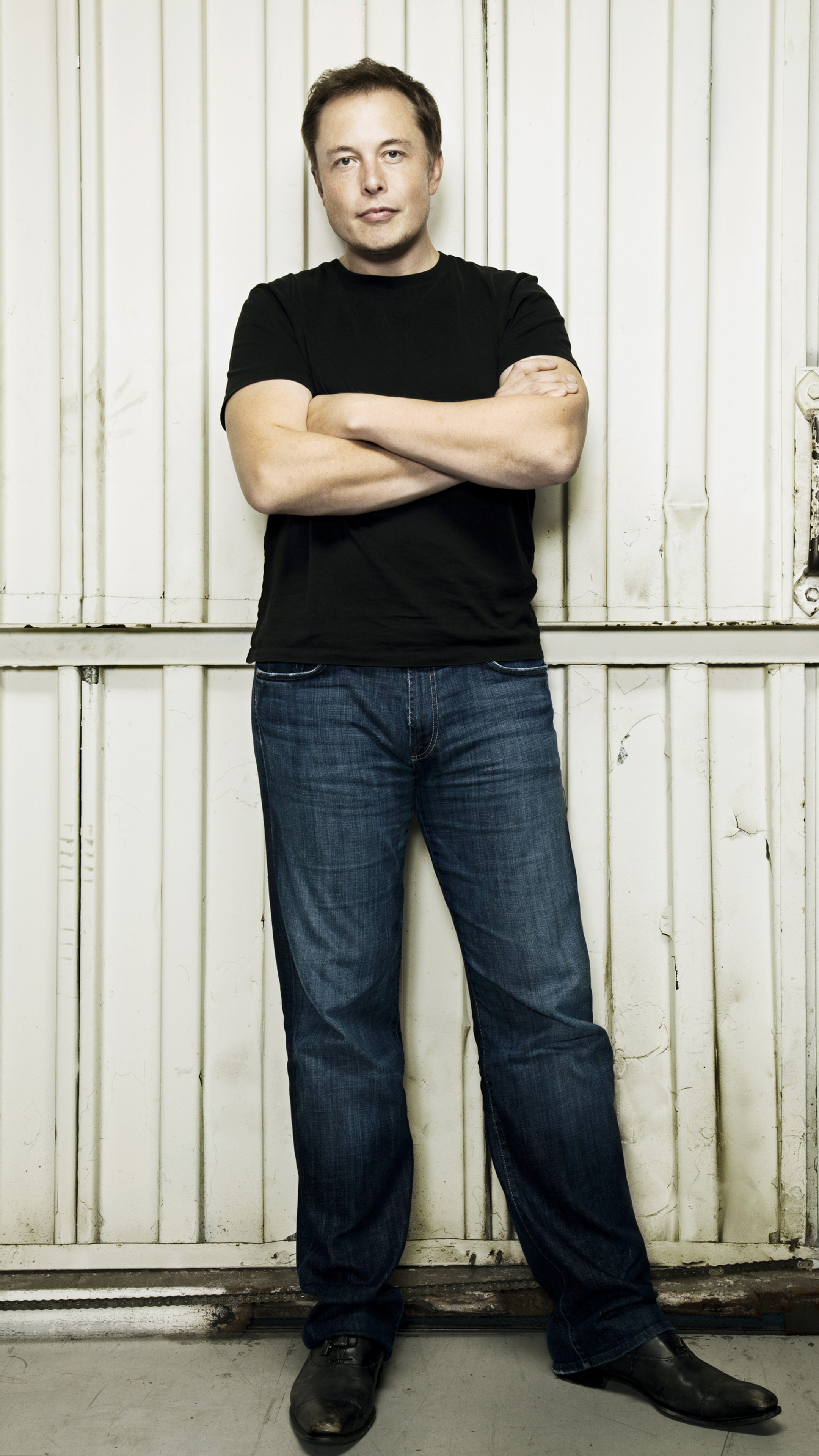 Elon Musk standing with arms crossed against a white wall