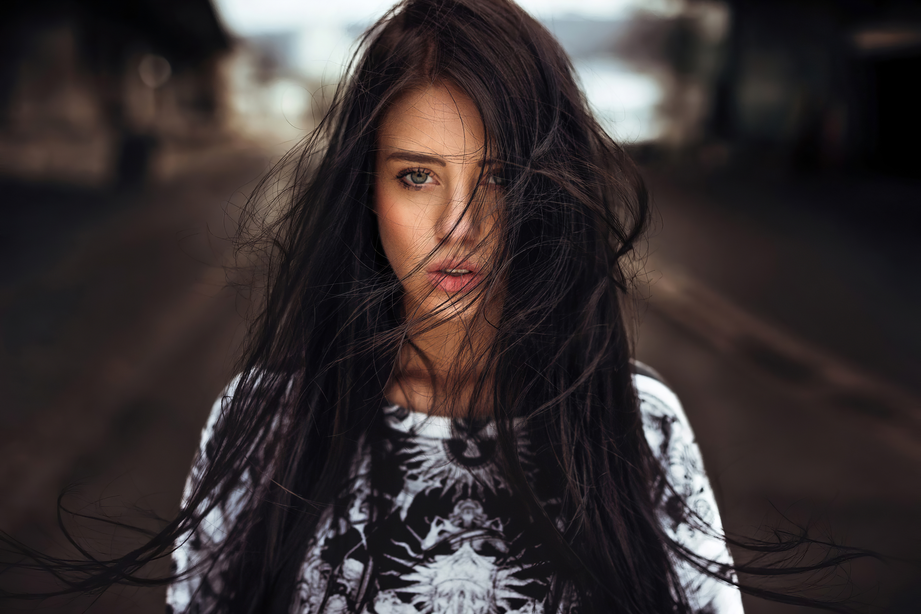 A young woman with long, dark hair blowing in the wind, wearing a graphic t-shirt, stands against a blurred background