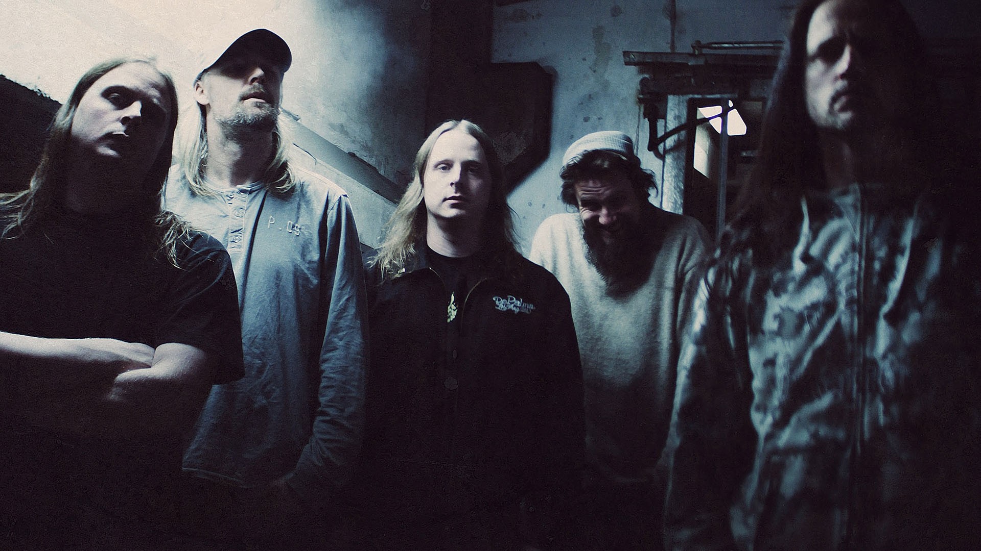 A dimly lit photograph of a band, likely a metal or post-rock group, featuring five men with long hair and casual attire