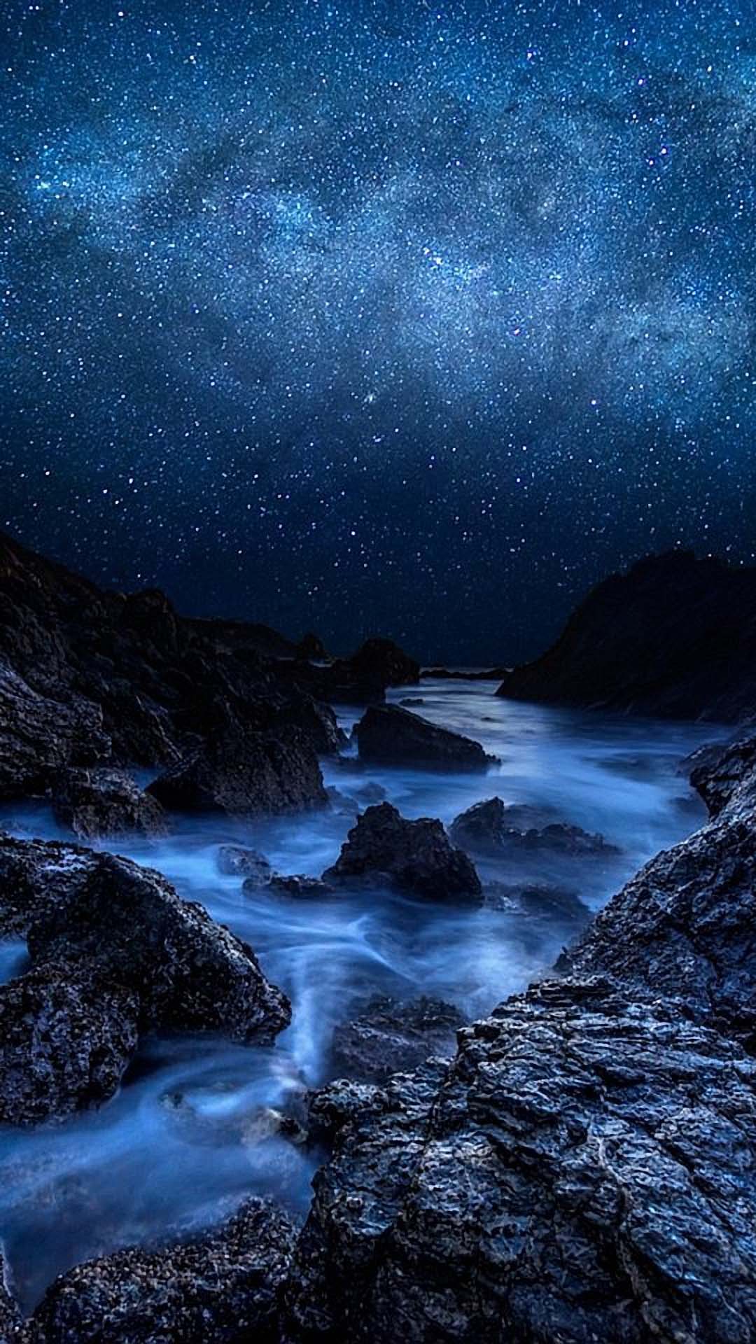 A breathtaking view of the Milky Way galaxy stretching across a night sky above a rocky coastline. The long exposure creates a smooth, ethereal effect on the water flowing between the dark rocks