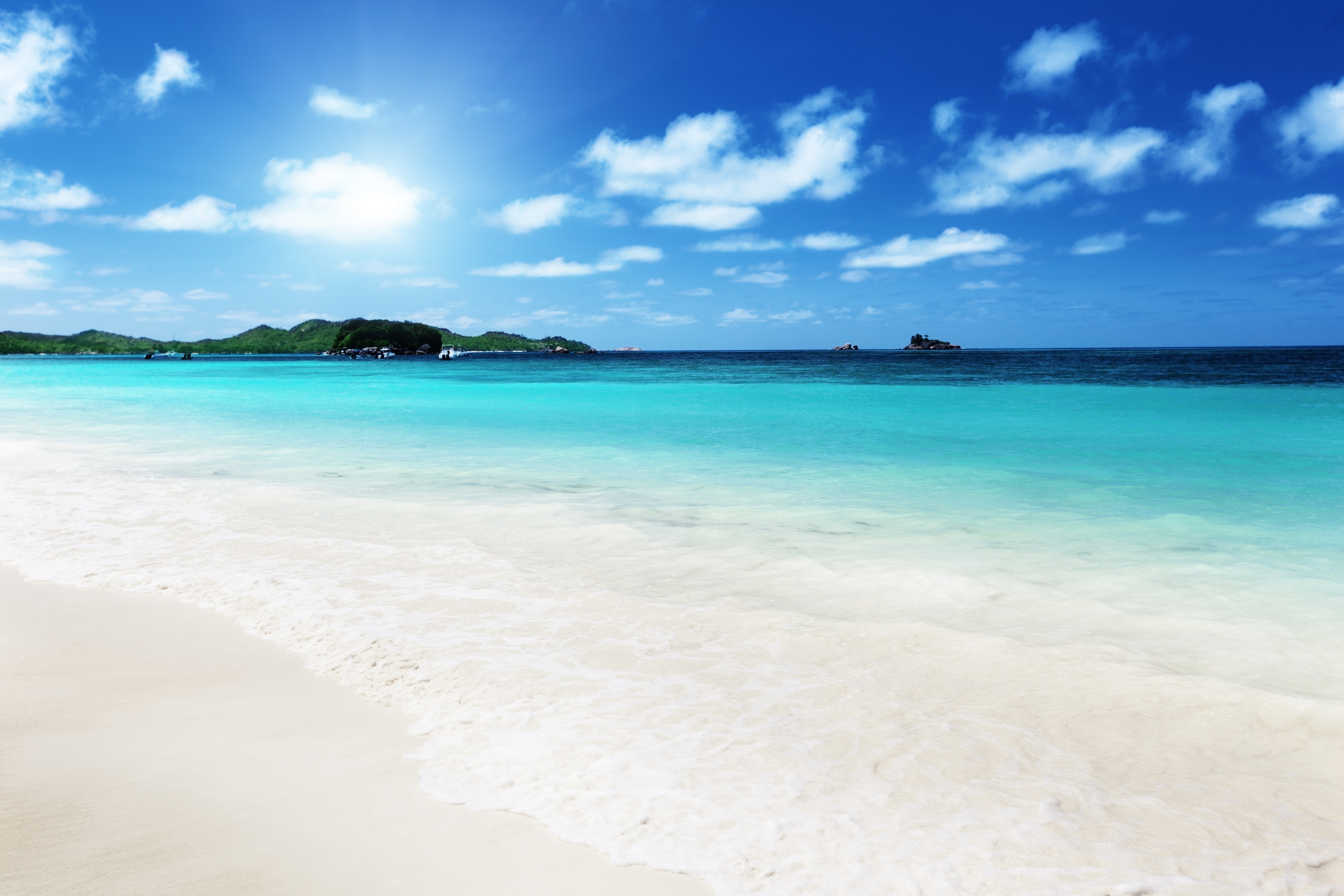 A breathtaking 4K beach wallpaper with pristine white sand, crystal-clear turquoise water, lush green islands, and a vibrant blue sky dotted with fluffy white clouds. The sun shines brightly, creating a perfect tropical paradise scene