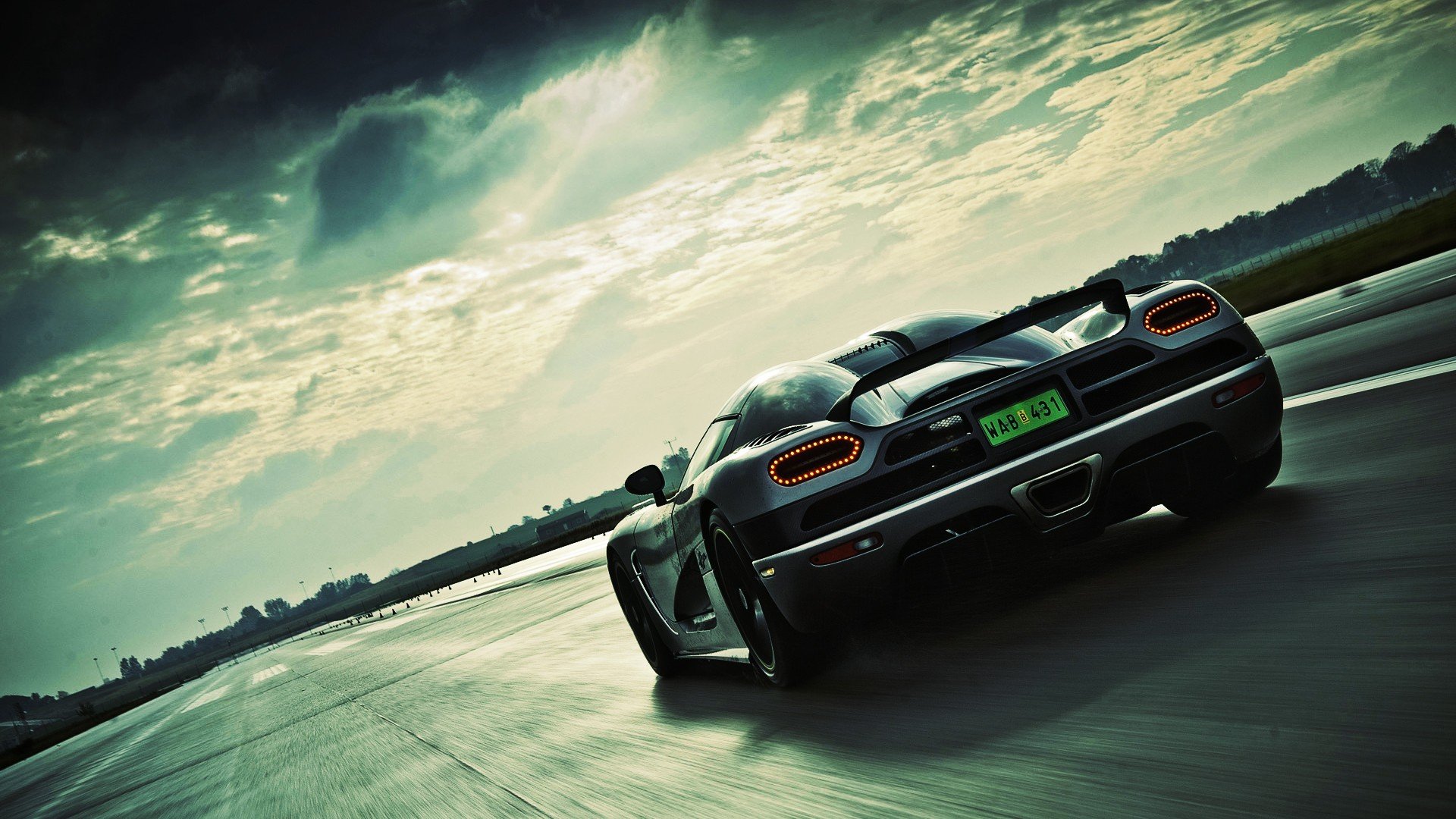 A silver Koenigsegg Agera speeds down a paved road under a cloudy sky