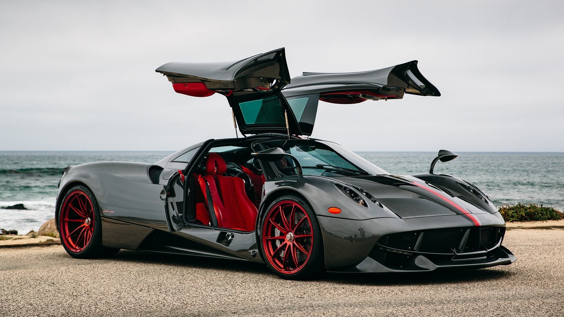 A dark gray Pagani Huayra with red accents and open dihedral doors parked by the ocean