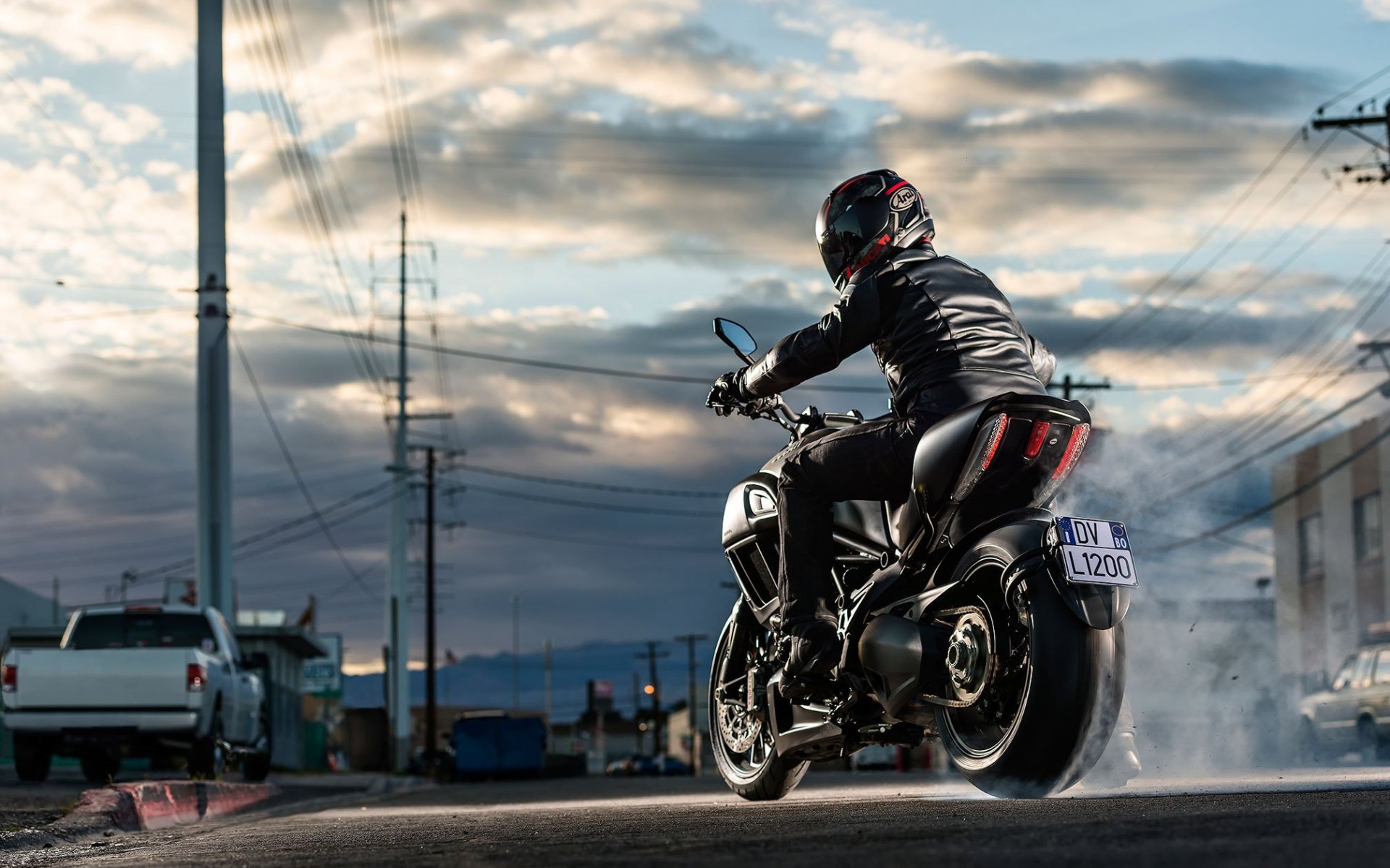A motorcyclist performs a burnout on a Ducati Diavel motorcycle, creating a cloud of smoke. The rider wears a black leather jacket and helmet