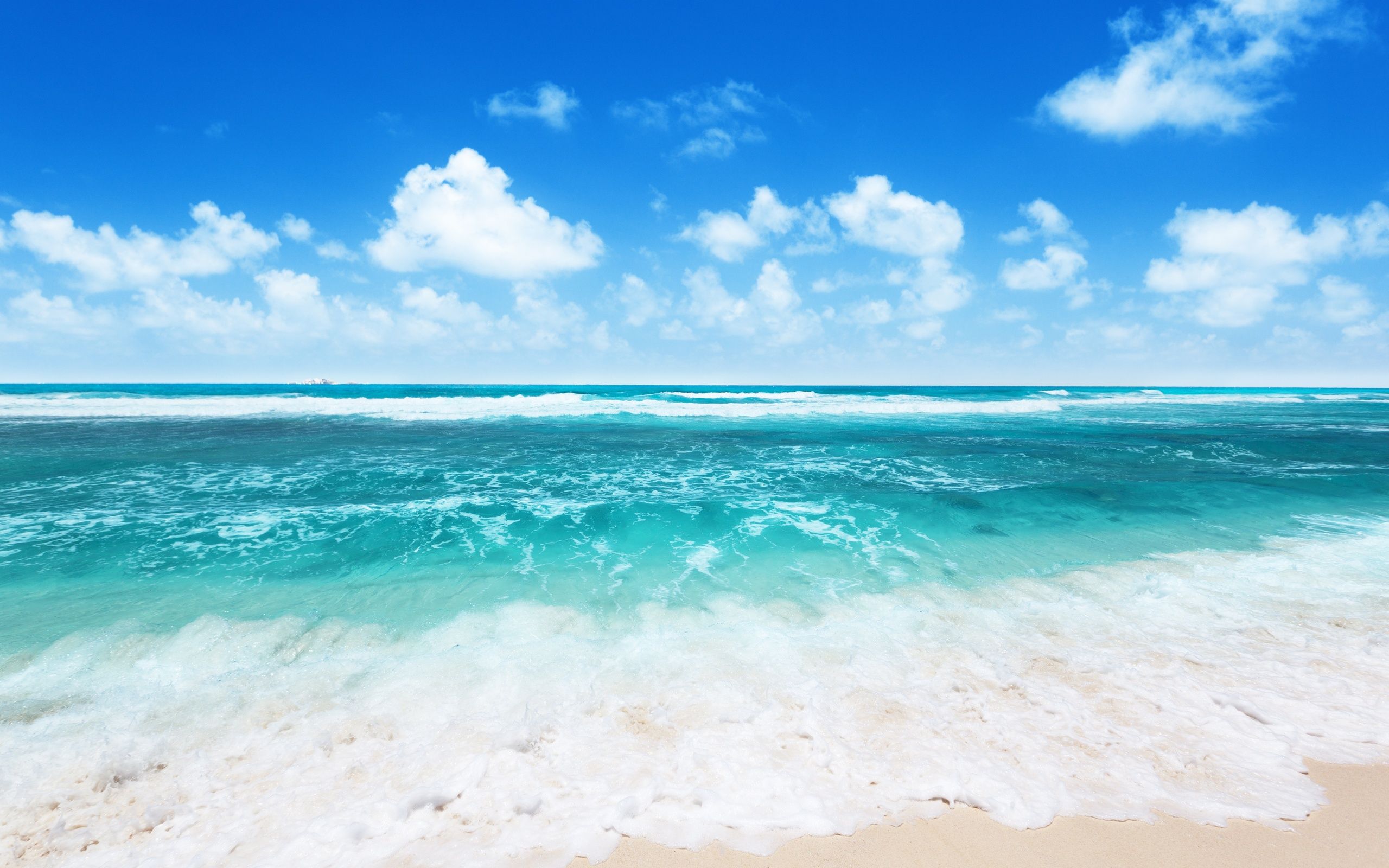 Turquoise ocean waves gently lapping onto a white sandy beach under a vibrant blue sky dotted with fluffy white clouds