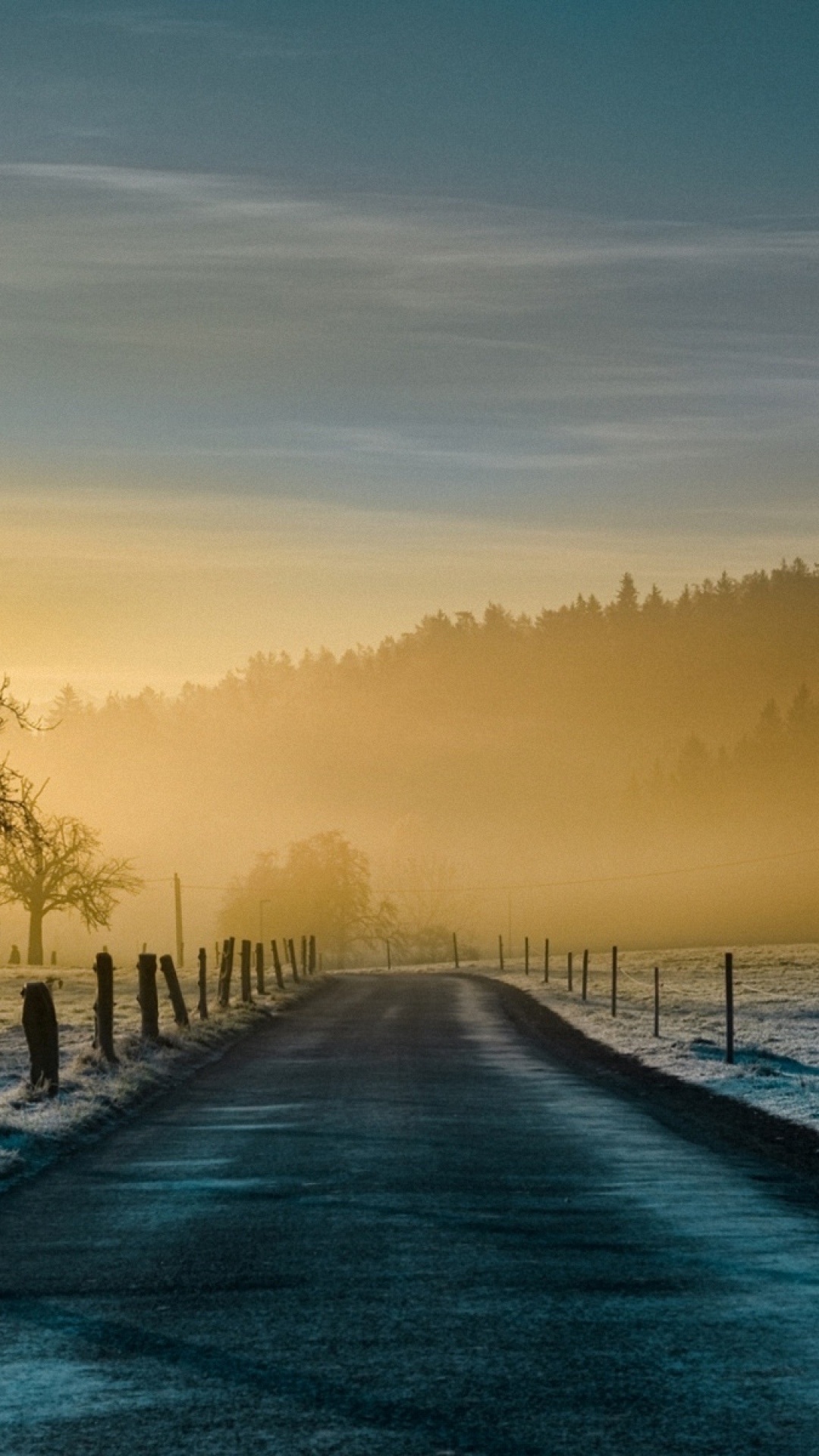A serene phone wallpaper featuring a misty sunrise over a country road, lined with frosted fields and a distant treeline