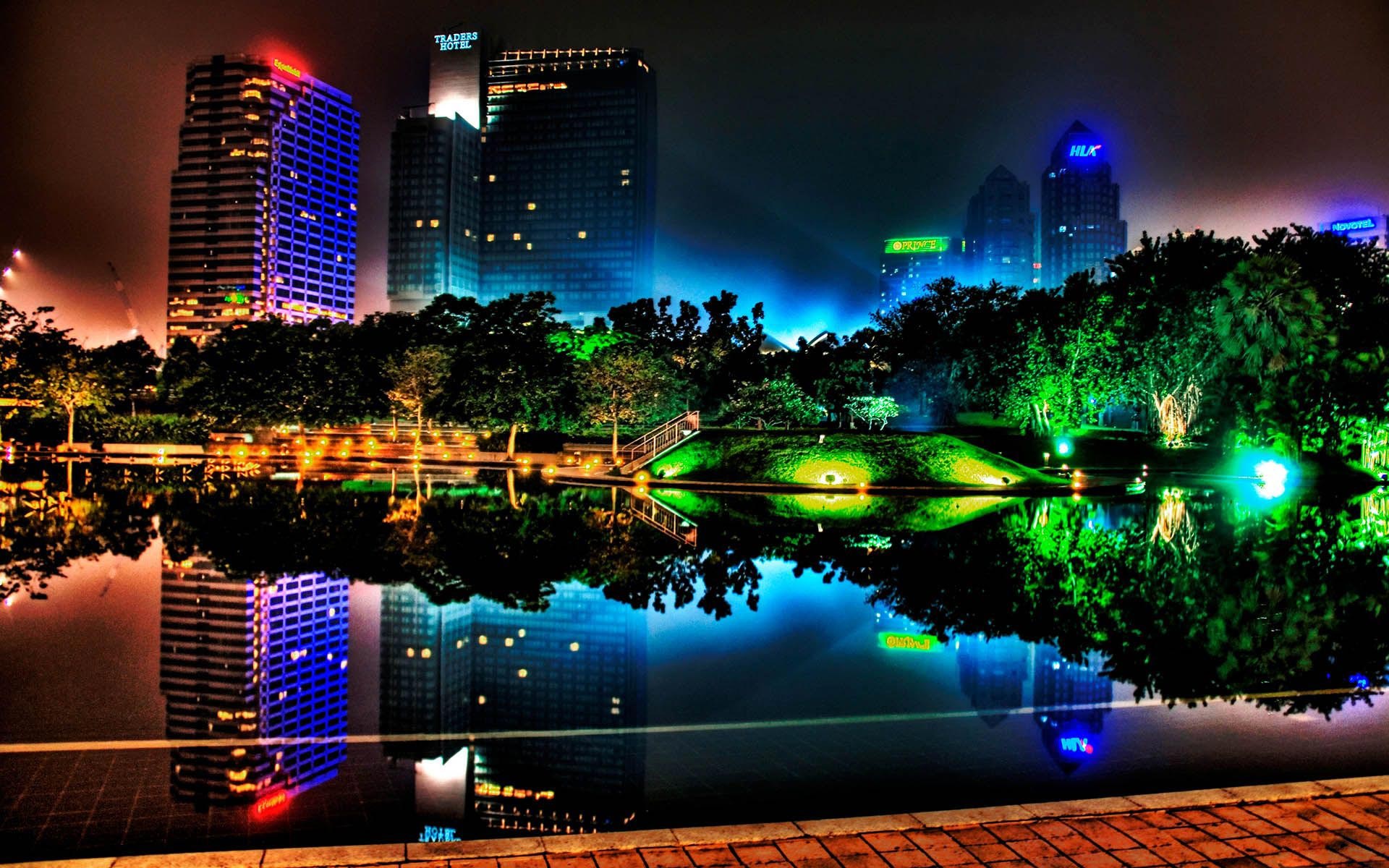 A vibrant cityscape at night, reflected in a tranquil pool. Colorful lights illuminate the buildings and trees, creating a stunning urban nightscape