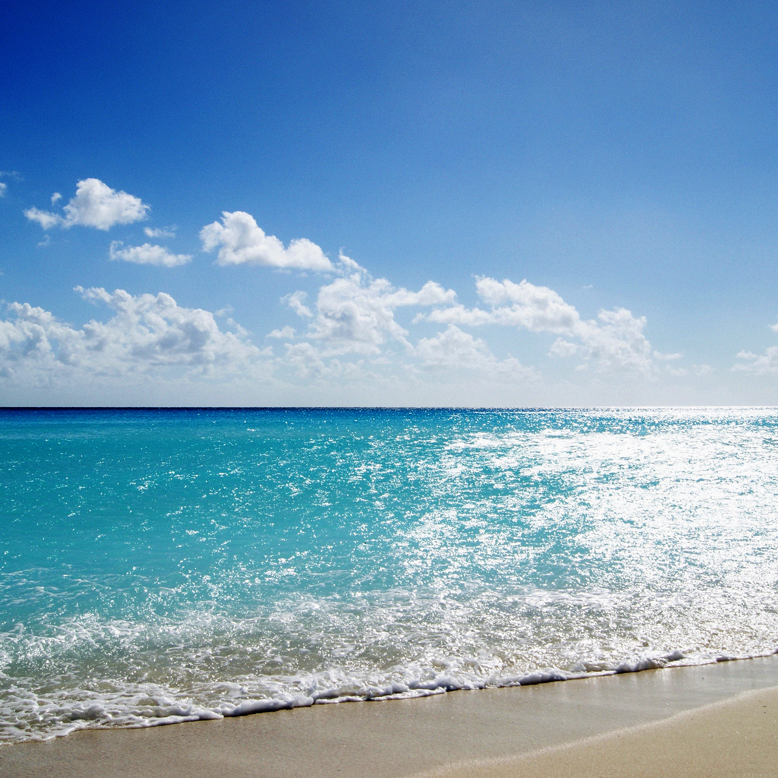 Turquoise ocean waves gently lapping against a sandy beach under a bright blue sky dotted with fluffy white clouds