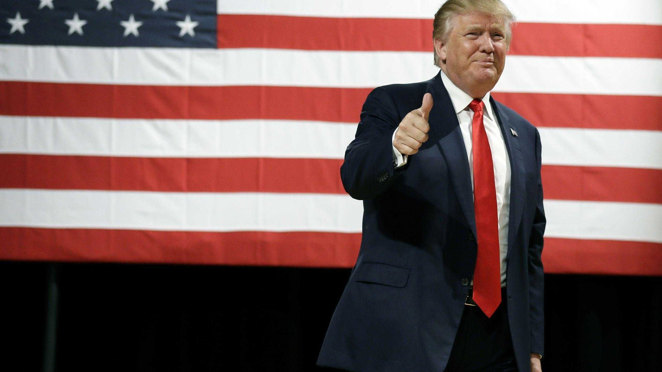 Donald Trump giving a thumbs-up gesture in front of an American flag, possibly during a 2024 campaign event
