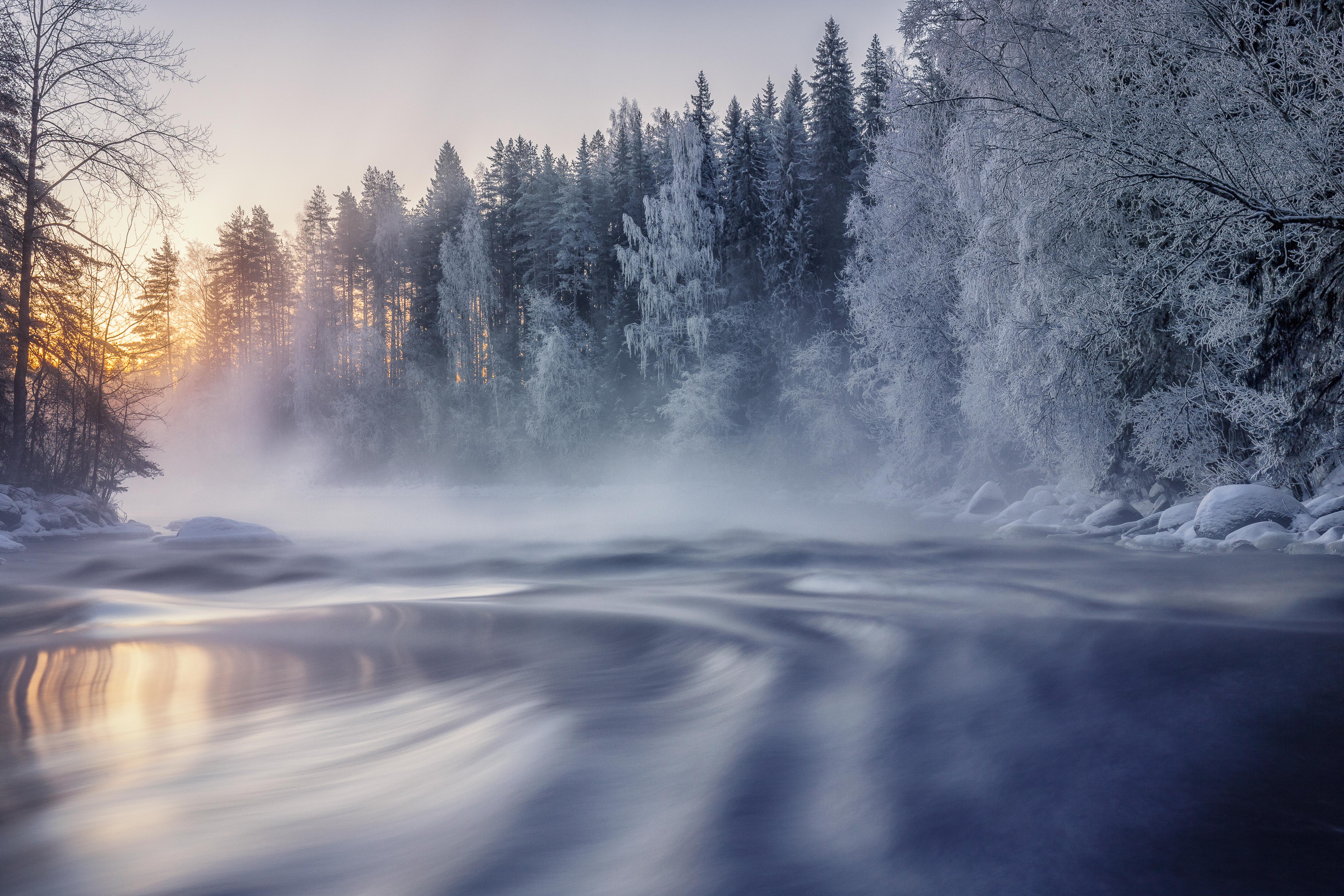 A breathtaking 4k winter wallpaper featuring a serene river flowing through a snow-covered forest at dawn. The mist rising from the water adds a touch of magic to the scene