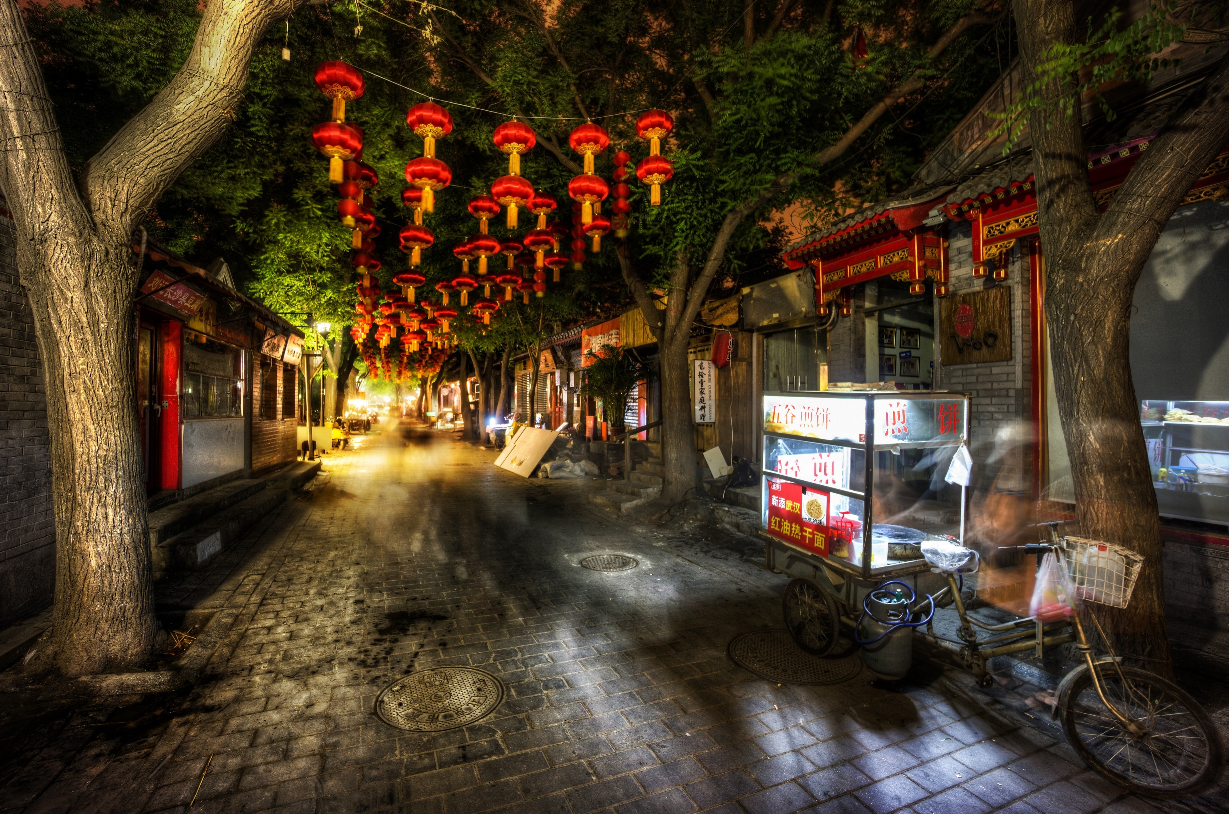 A charming narrow street in a Chinese town at night, illuminated by the warm glow of red lanterns and a street food vendor's stall. The cobblestone street and traditional architecture evoke a sense of history and culture
