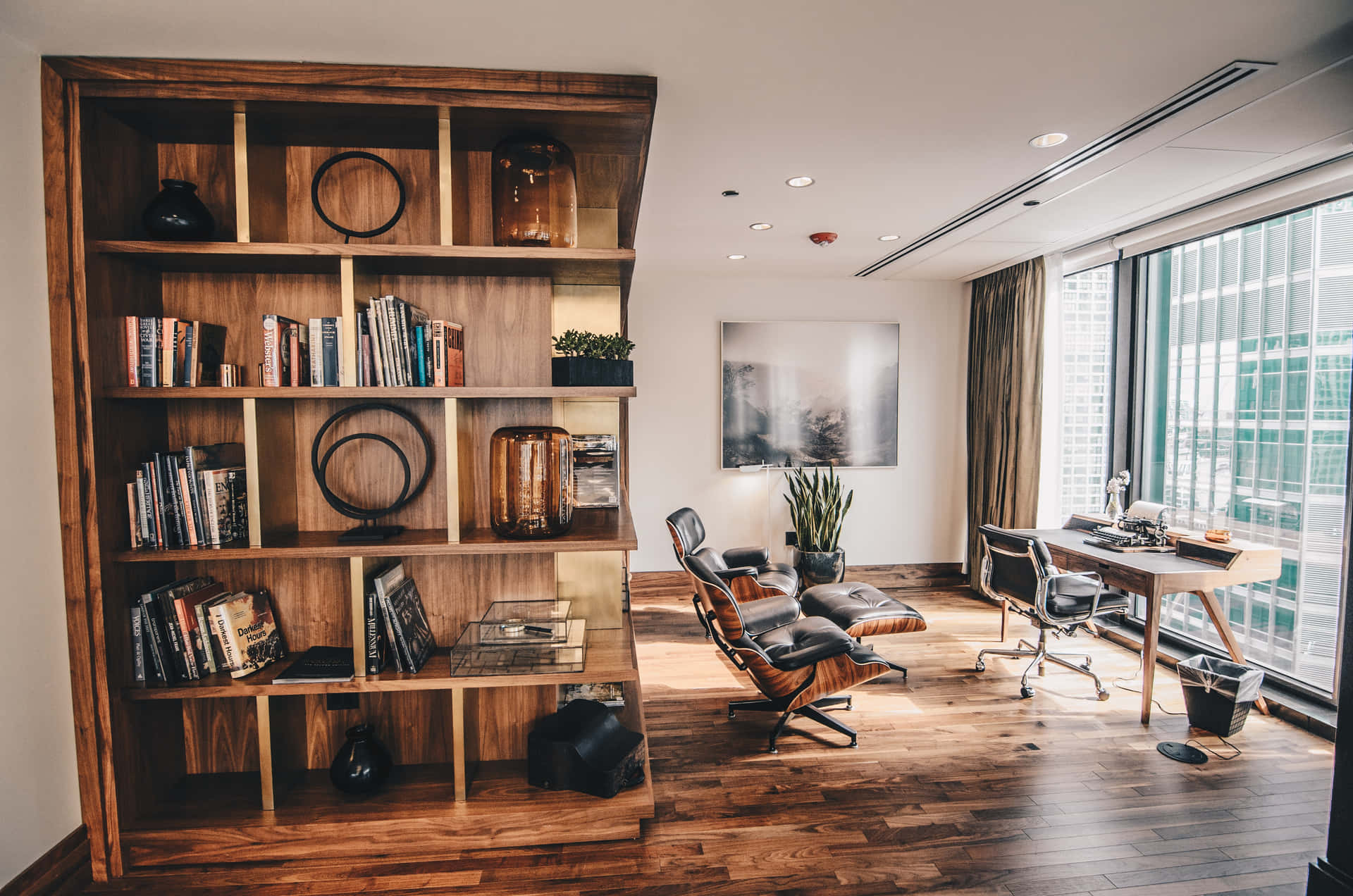Modern home office with a stylish wooden bookshelf, two lounge chairs, and a desk by the window overlooking the city