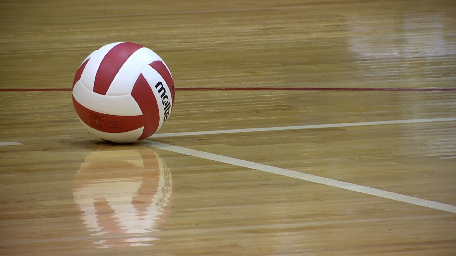 A volleyball sits on a polished wooden court, resting near the intersection of boundary lines. Its reflection gleams on the floor's surface