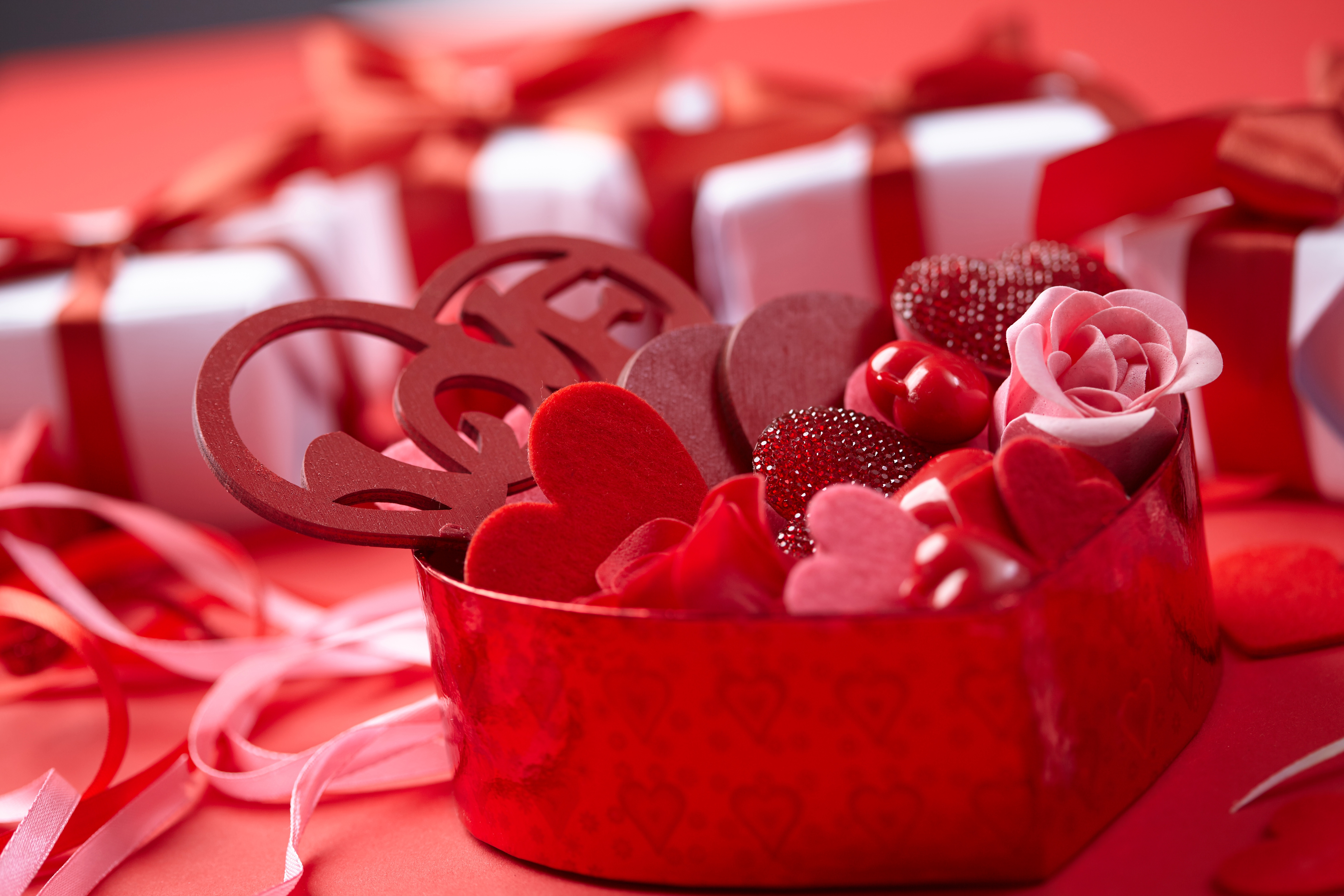 A close-up of a heart-shaped box filled with Valentine's Day treats, including candies, hearts, and a pink rose, set against a backdrop of wrapped gifts and ribbons in shades of red and white