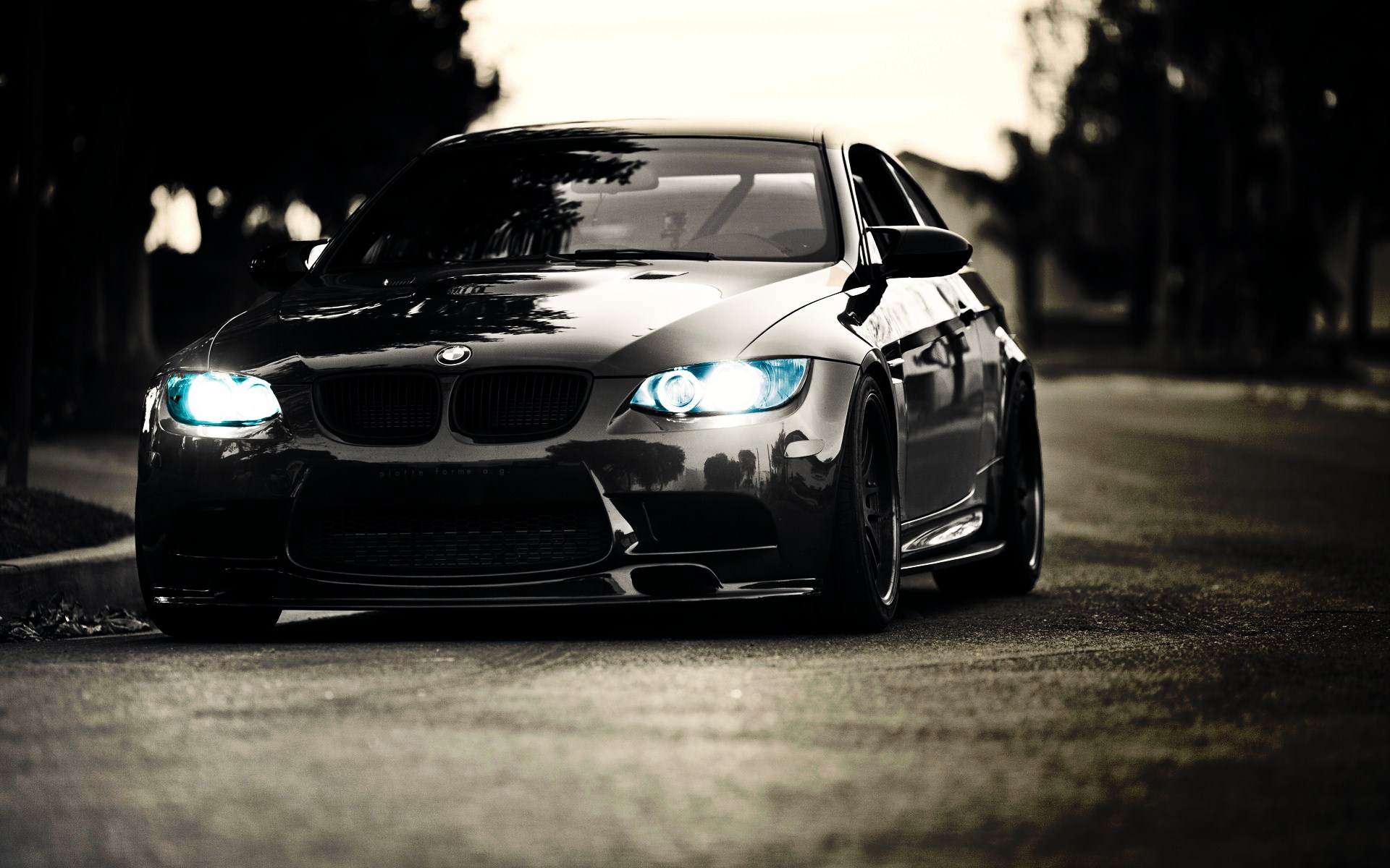 A sleek black BMW M3 coupe with its headlights on, parked on a dimly lit street