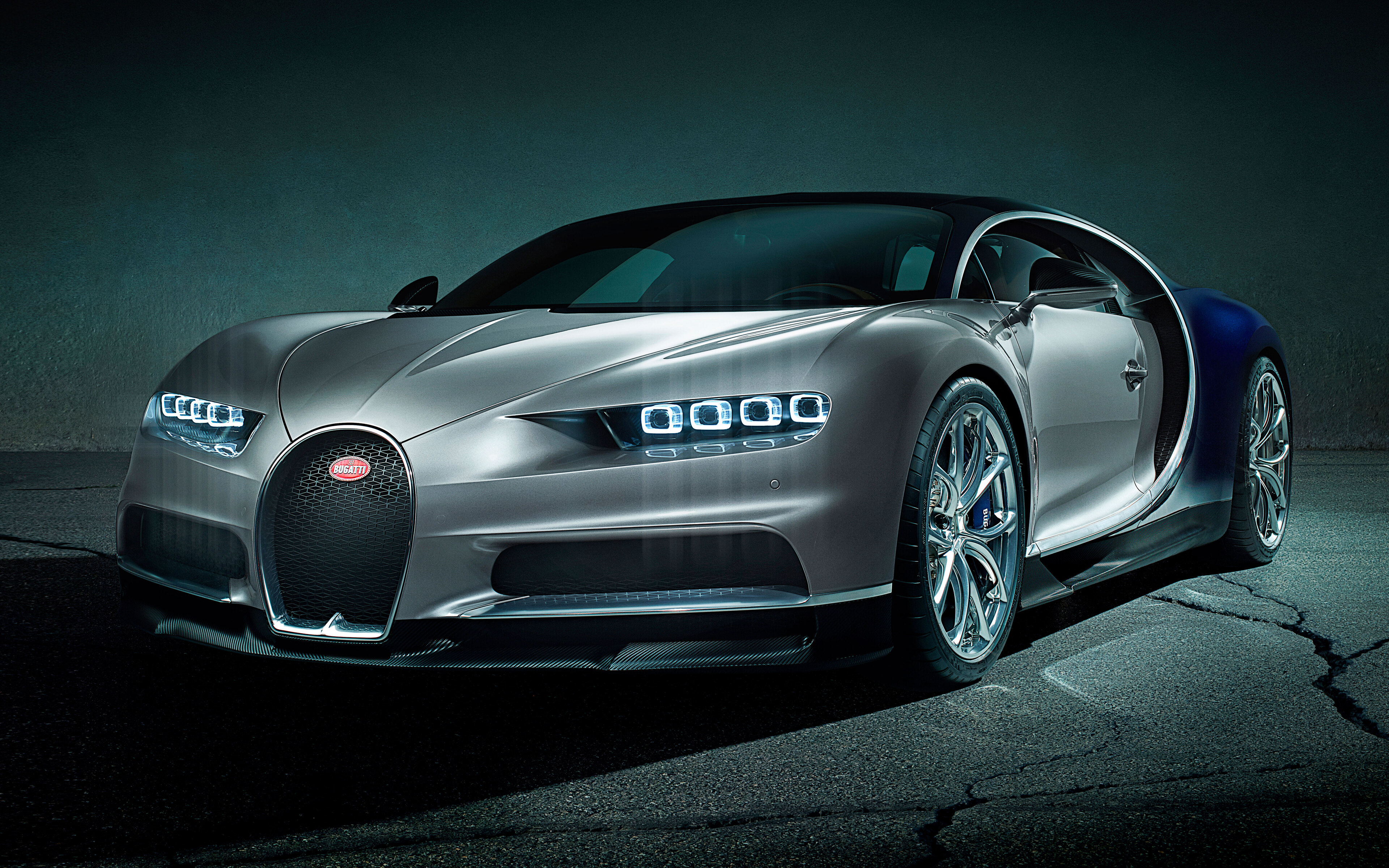 A silver and blue Bugatti Chiron sports car parked on a dark asphalt surface, illuminated by headlights, showcasing its sleek design and luxurious details