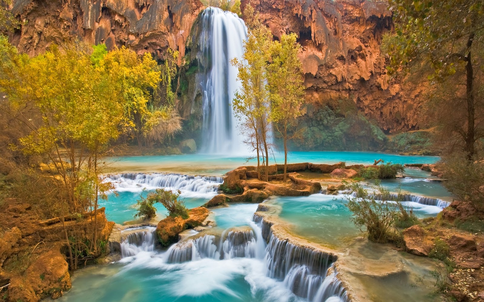 A breathtaking waterfall cascades down a red rock canyon into a turquoise pool, surrounded by vibrant autumn foliage