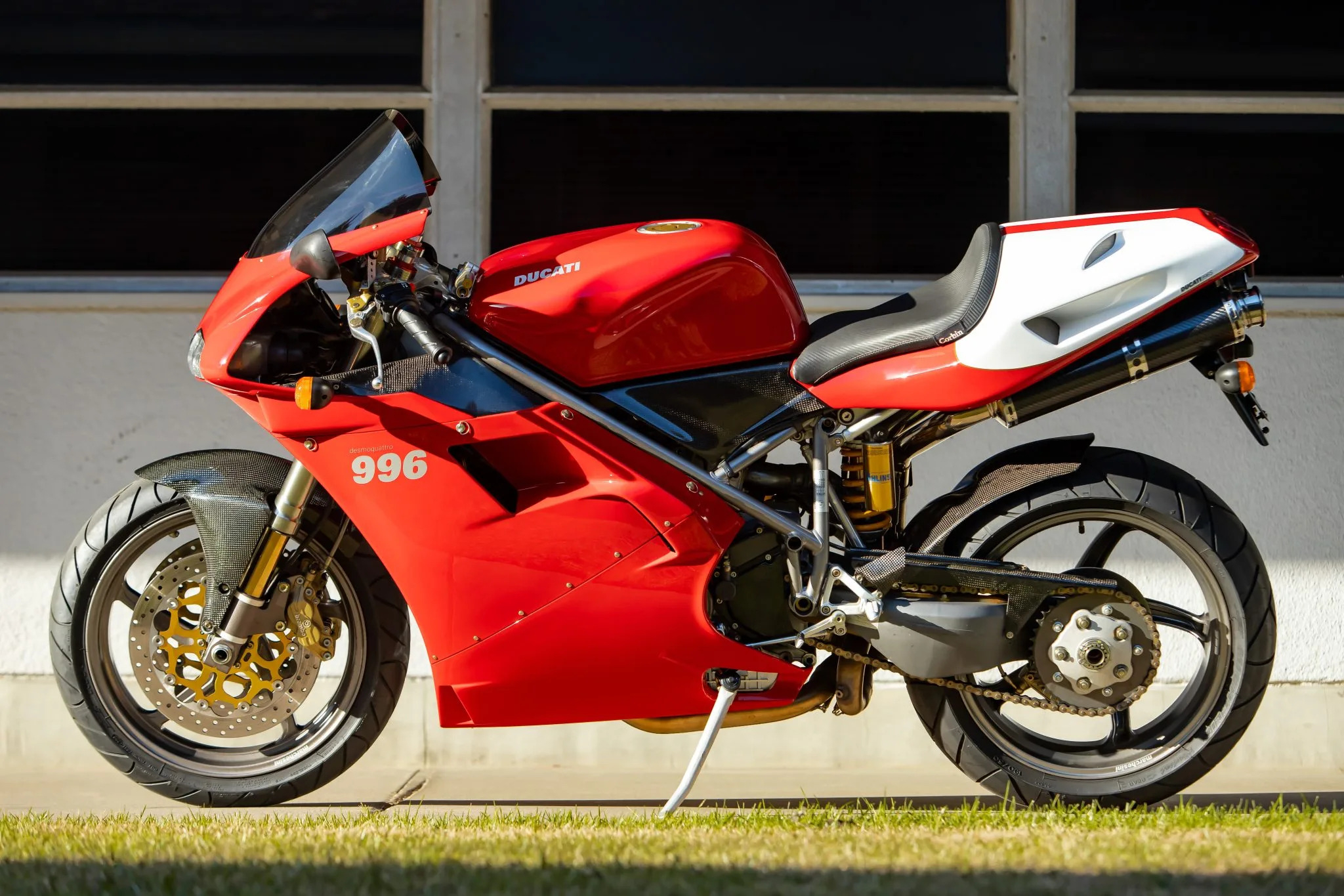 A Ducati 996 sportbike parked in front of a white wall with grass in the foreground. The bike is primarily red and white, highlighting its iconic design and racing heritage