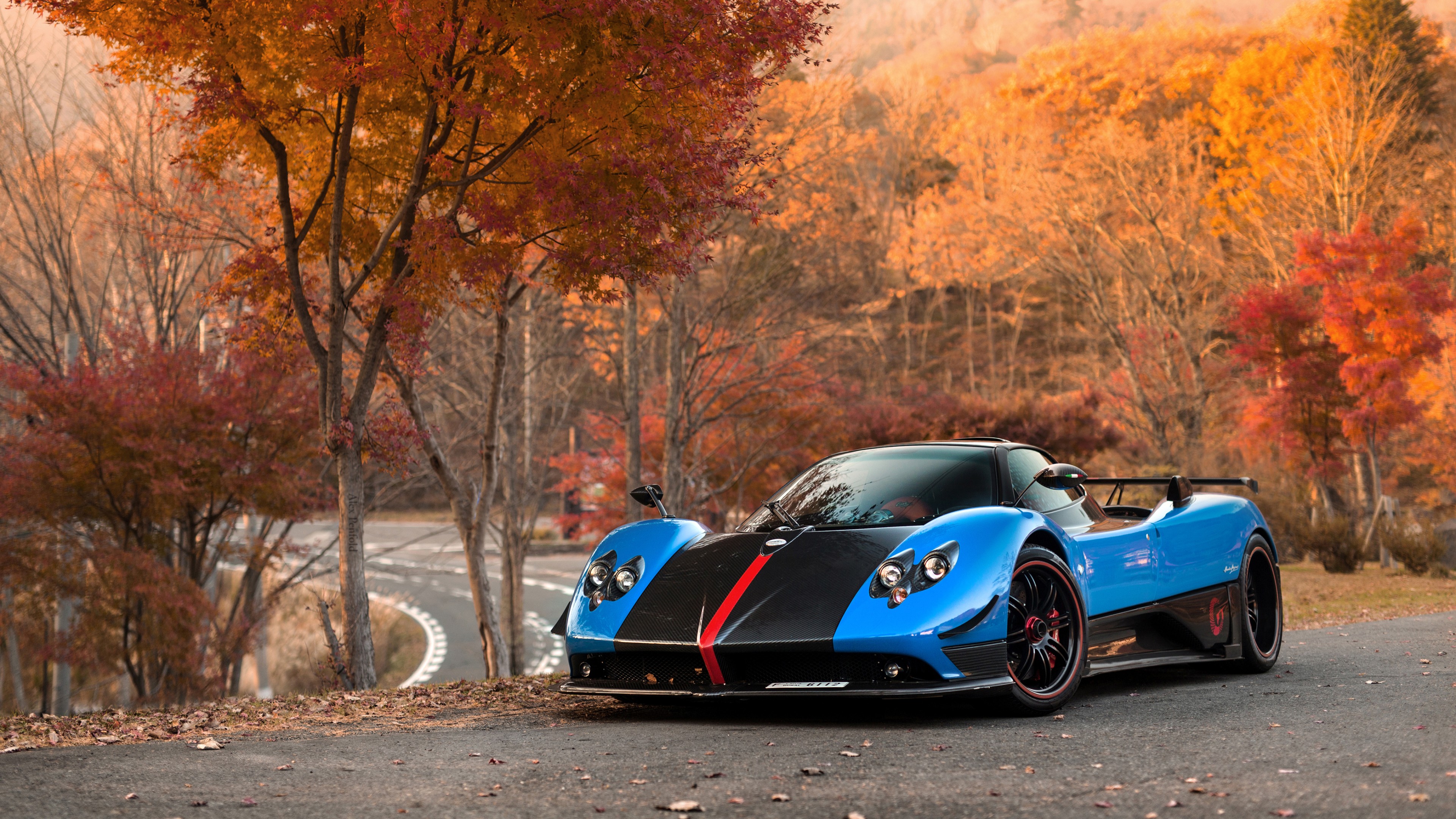 A vibrant blue Pagani Huayra supercar parked on a scenic road, framed by autumn foliage