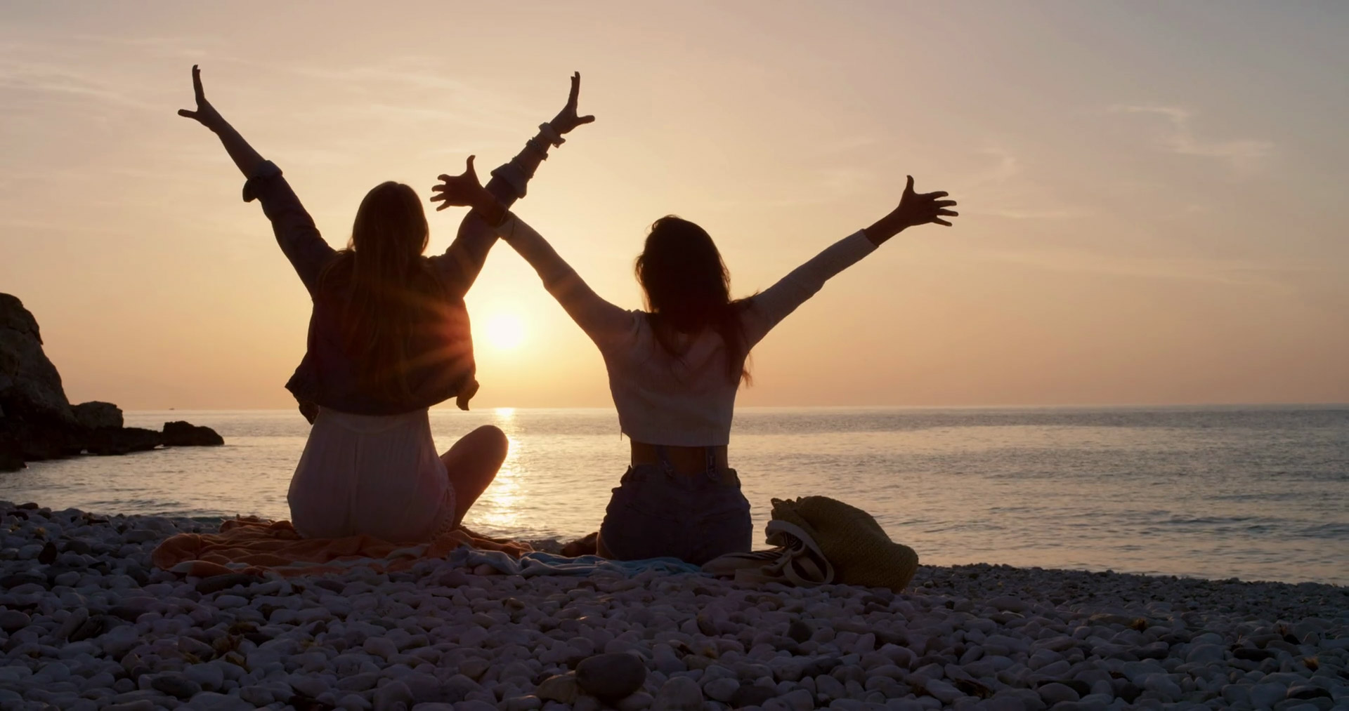 Two friends with arms outstretched enjoying a beautiful sunset on a pebble beach, embodying friendship and happiness
