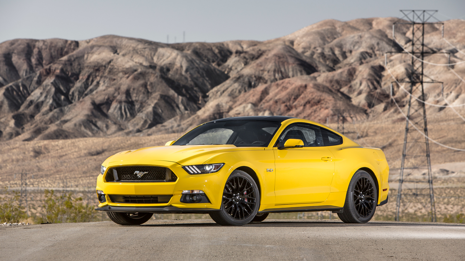 A bright yellow Ford Mustang sports car parked on a desert road, with rugged mountains in the background