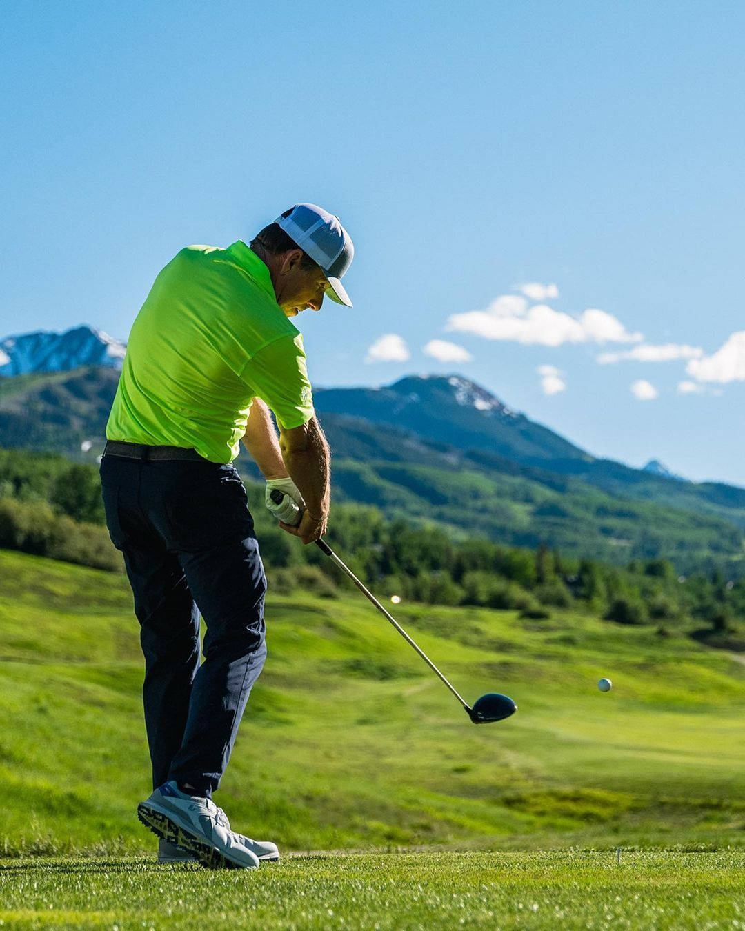 A golfer tees off on a lush green golf course against a backdrop of majestic mountains